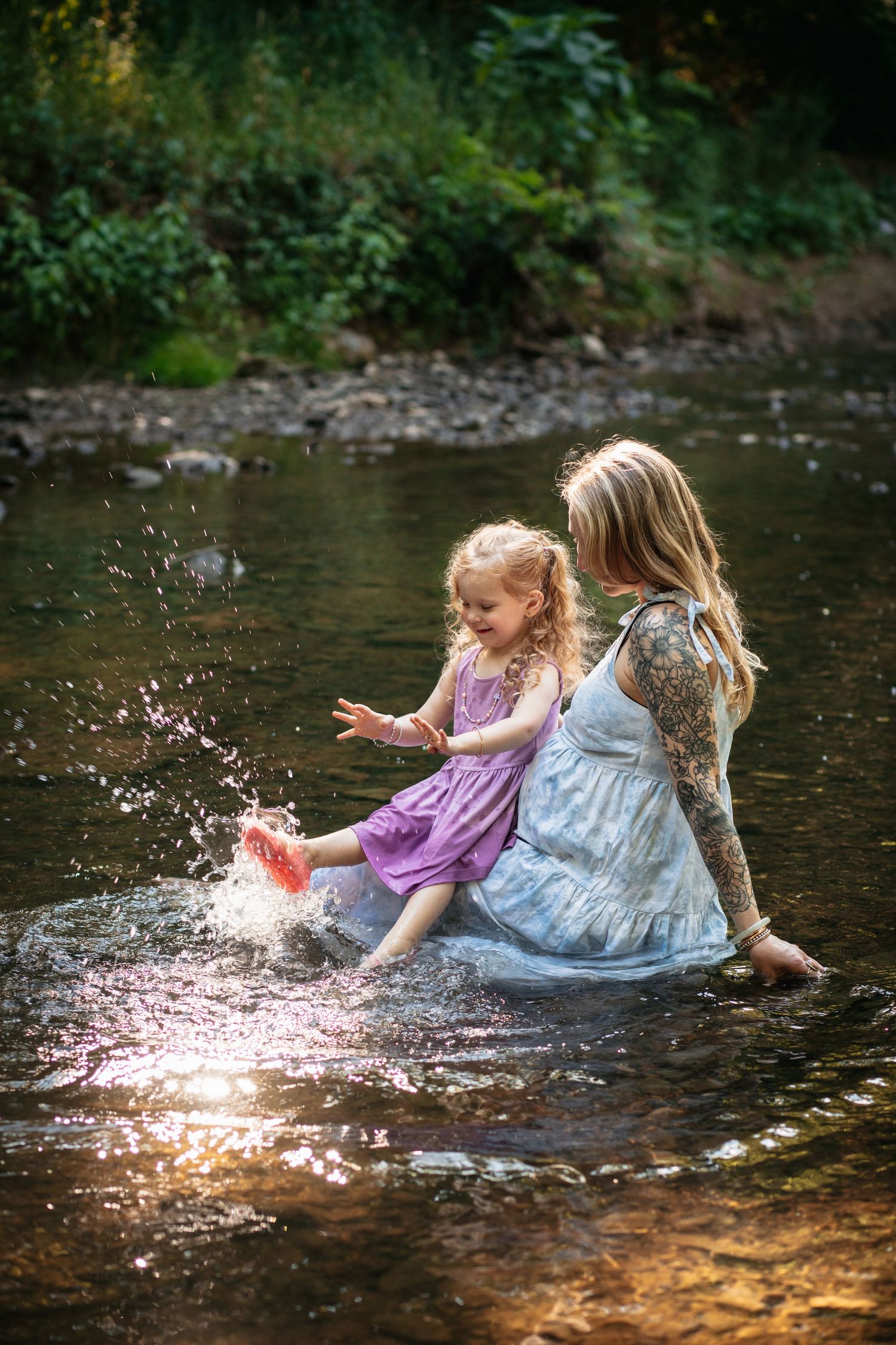 Two children in flowing dresses splash and play together in a shallow stream on a summer afternoon.