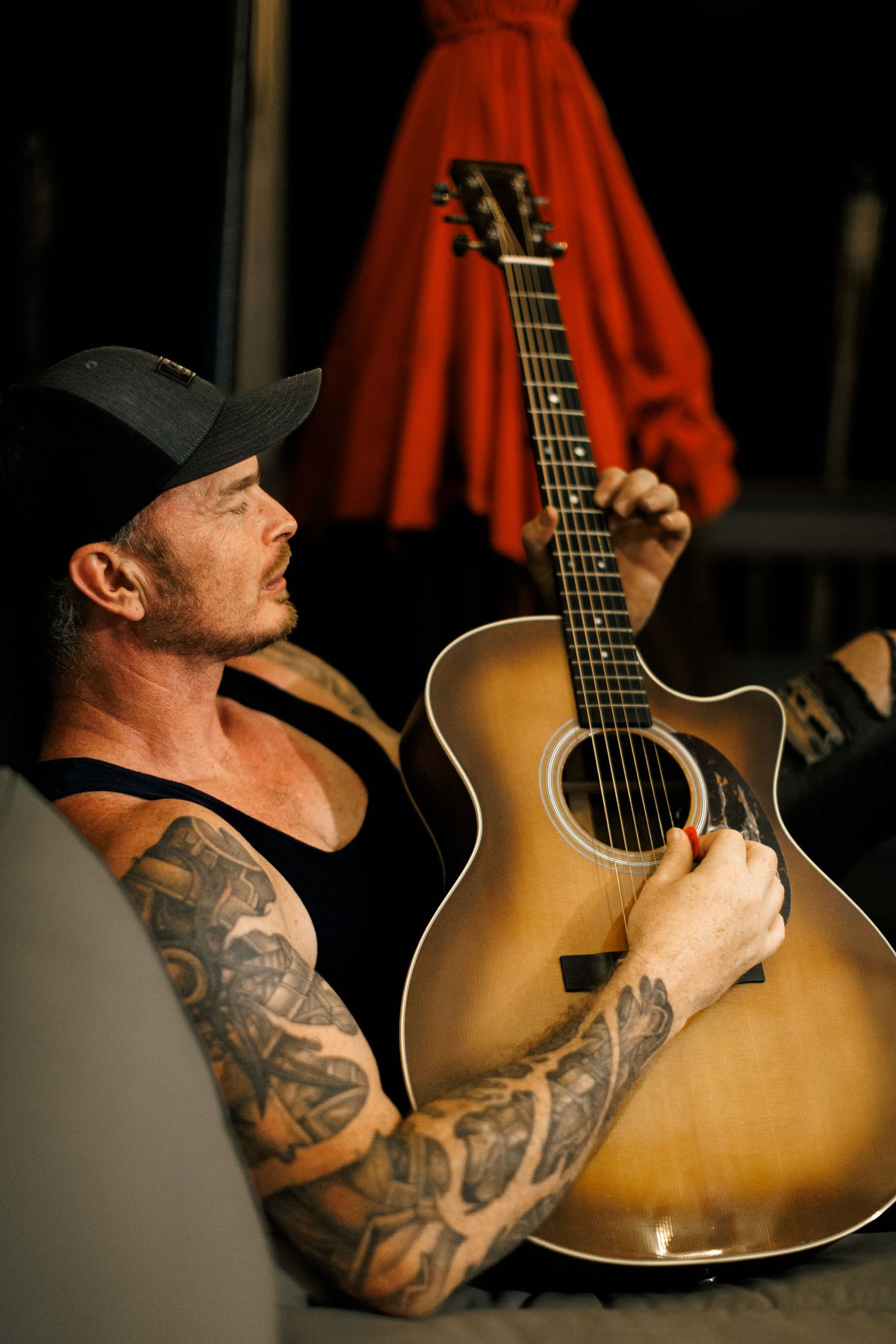 Musician with tattooed arms holds an acoustic guitar while performing in front of red stage curtains.