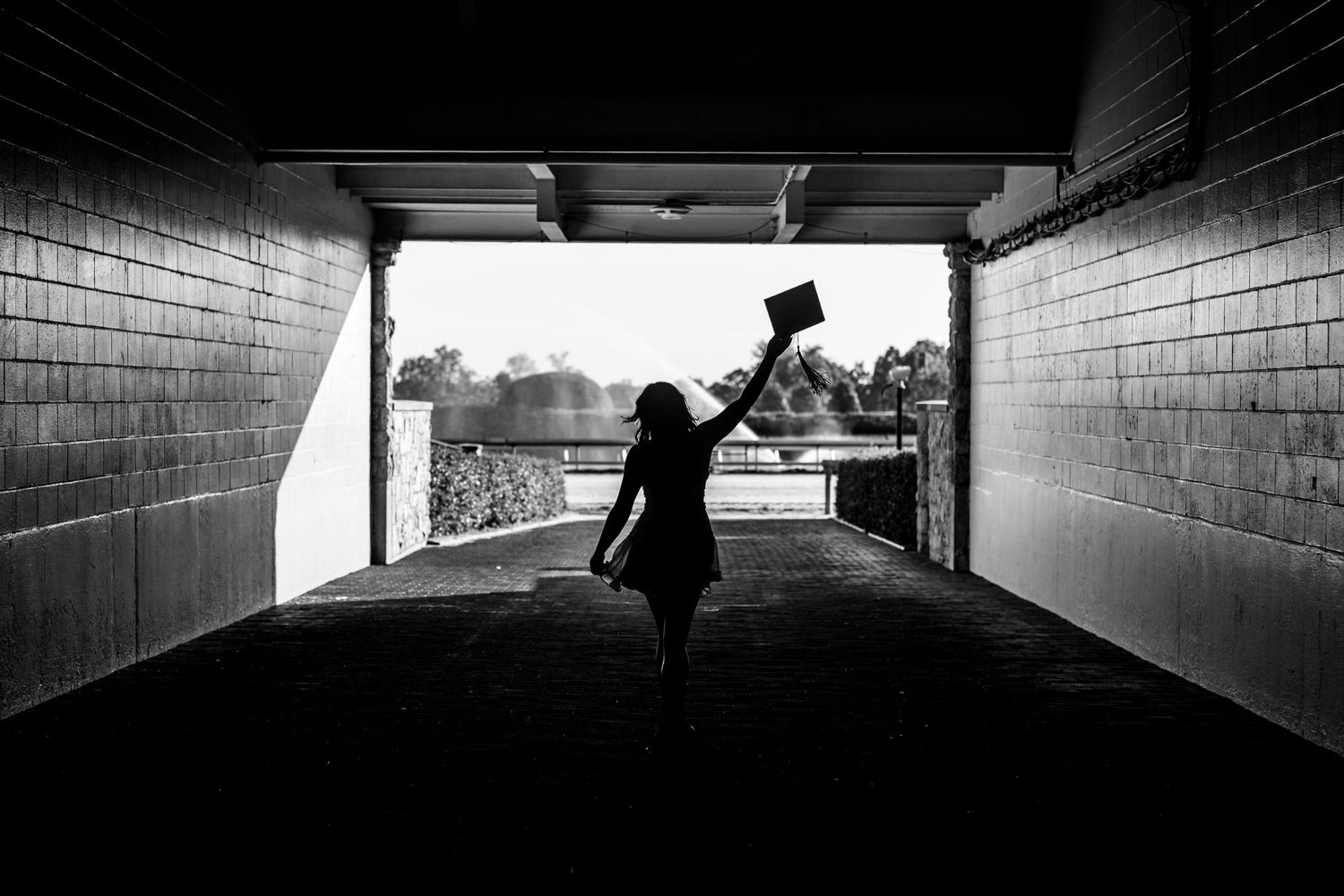 Silhouette in tunnel raises graduation cap triumphantly against bright background in black and white photograph.