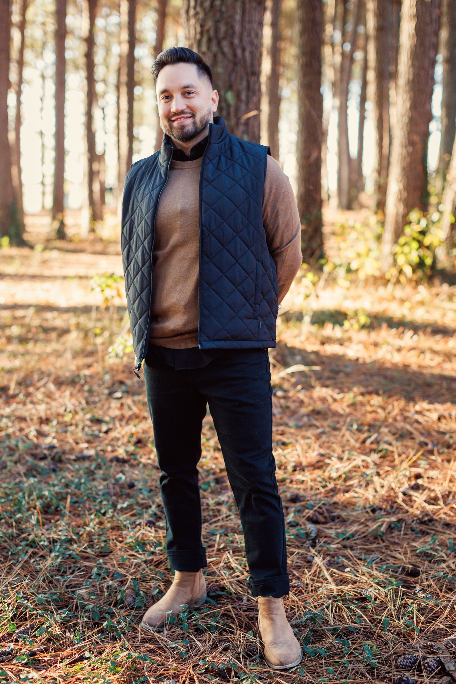 Person wearing brown sweater and navy quilted vest stands in autumn forest with fallen pine needles on ground.