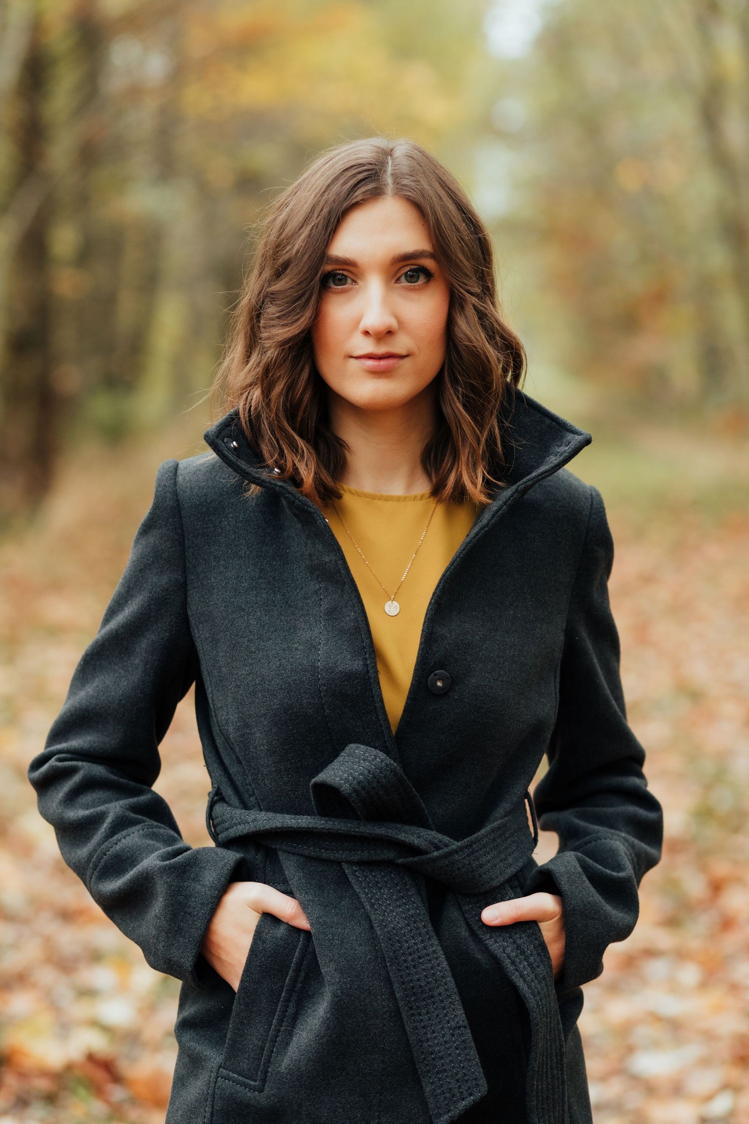 Fall fashion portrait showcasing a black belted coat and yellow top among autumn leaves.