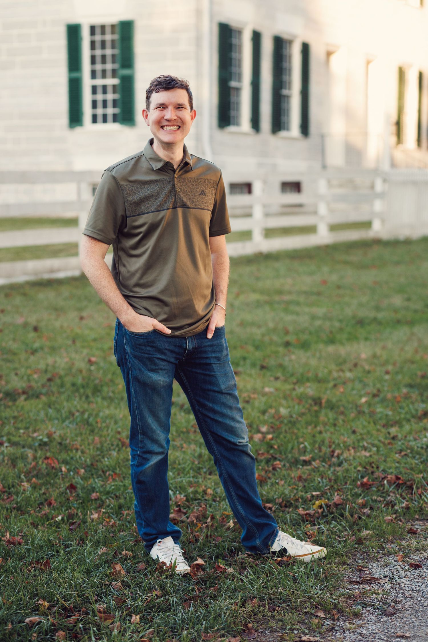 Person standing on grass wearing olive polo shirt and jeans in front of white building with green shutters.