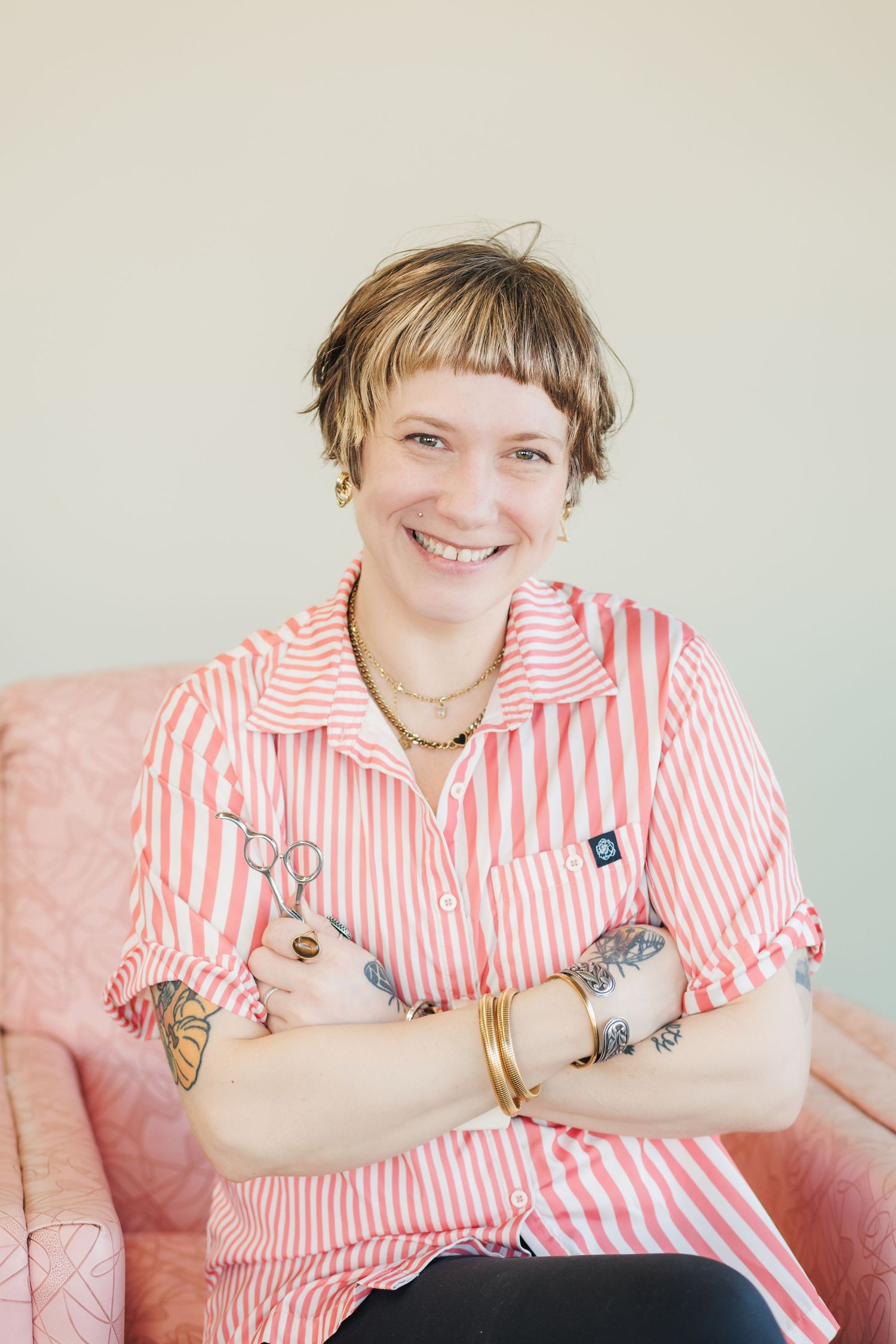 Person in pink and white striped shirt sitting on pink couch with crossed arms and visible tattoos.