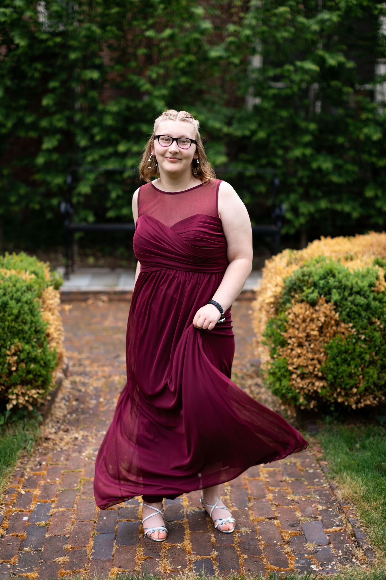 Woman in burgundy formal dress twirls on brick pathway surrounded by autumn foliage.