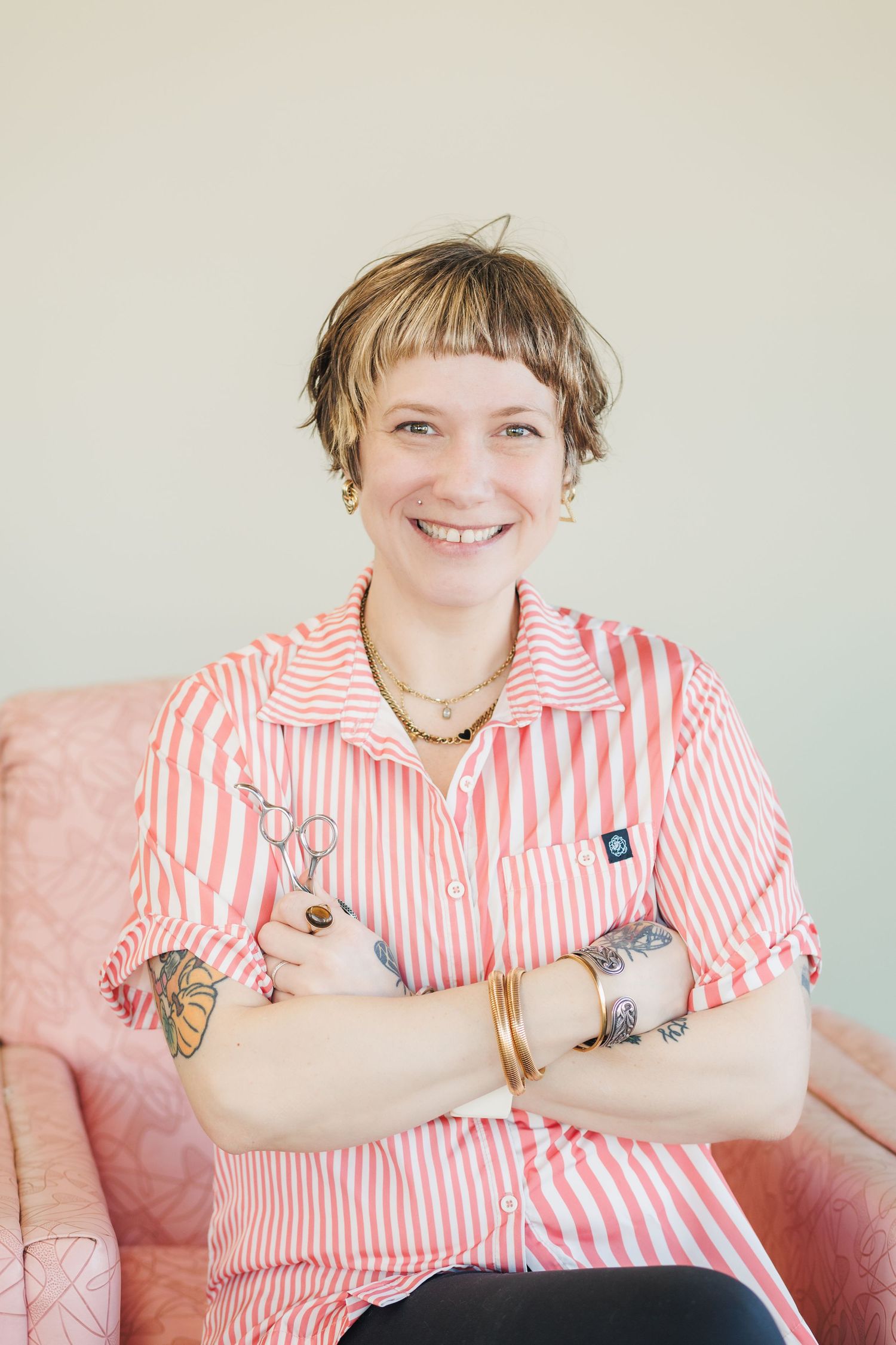 Person wearing pink and white striped shirt with tattoos sits smiling in pink chair against light background.