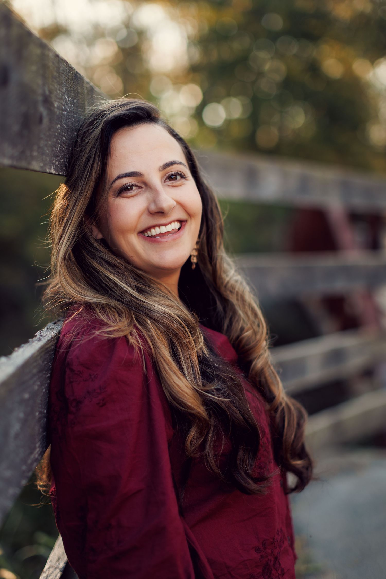 A person in a burgundy sweater smiles warmly while sitting outdoors with soft bokeh lighting in the background.