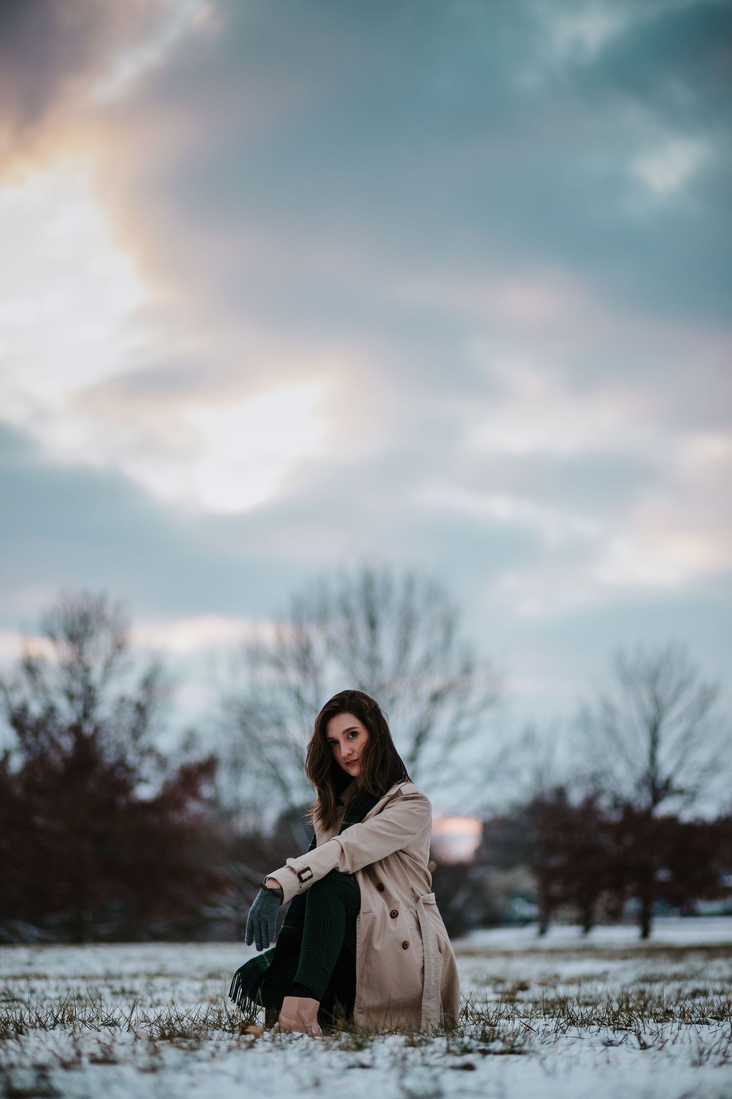Artistic winter portrait in beige coat against snowy landscape with dramatic sky.