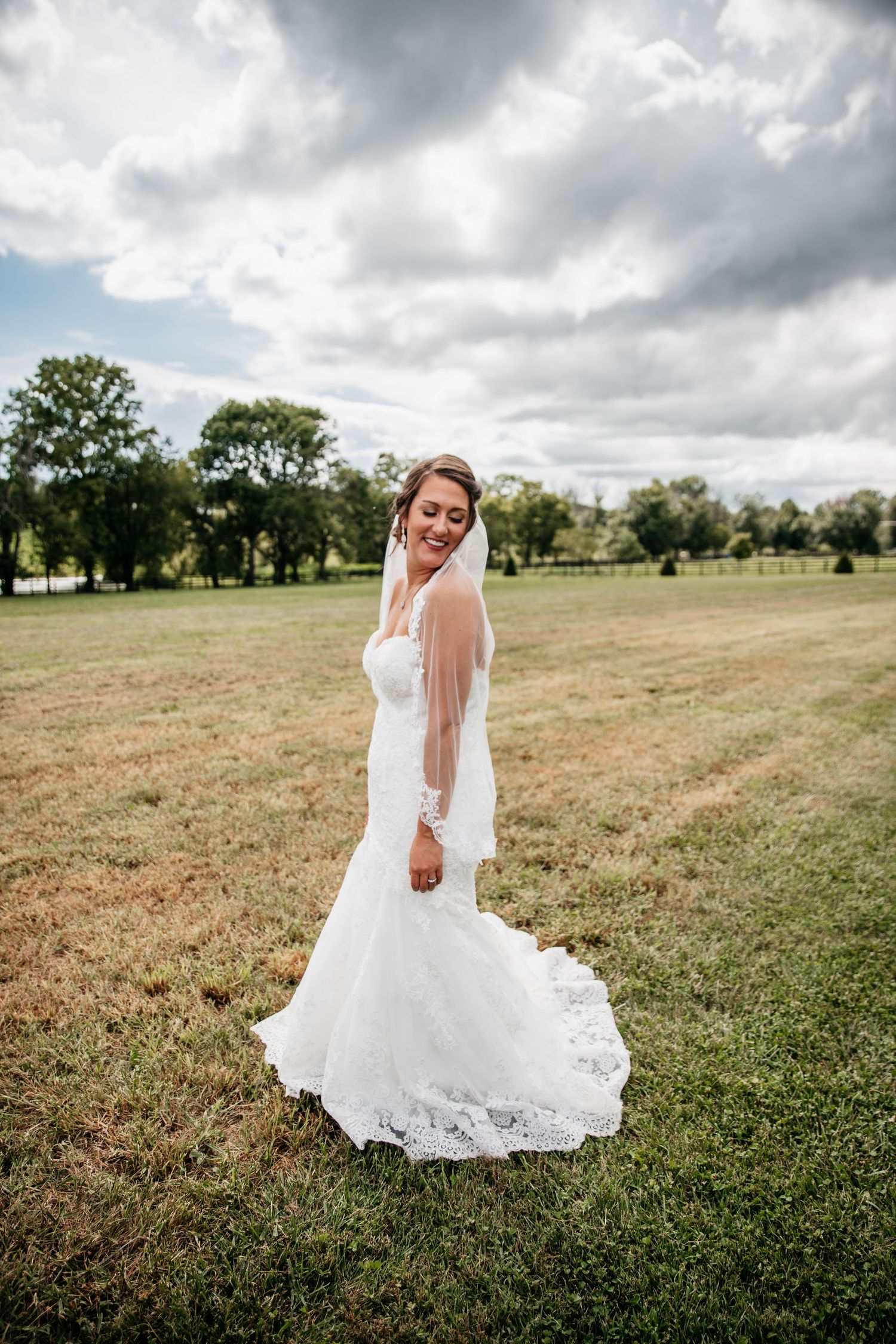 Elegant bride in flowing white dress poses in a grassy field under dramatic sky.