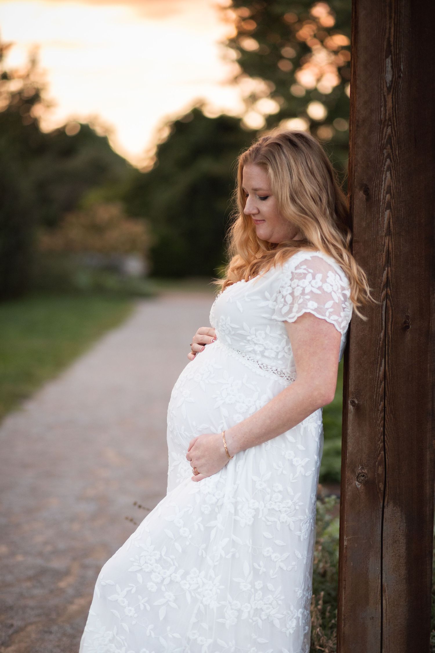 Pregnant woman in white lace dress poses by wooden post during sunset maternity session.