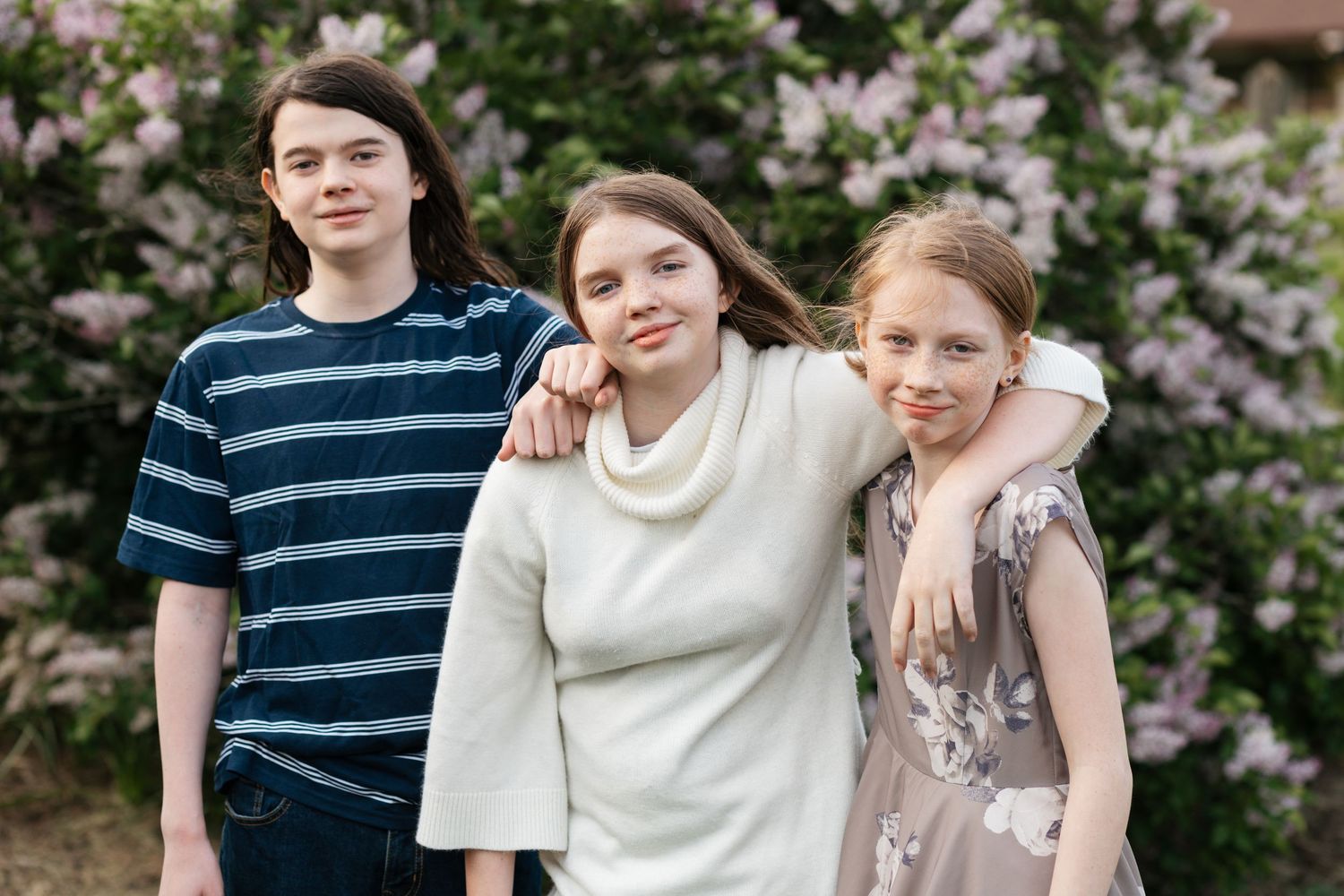 Three young people in casual attire stand together smiling in front of blooming pink flowers on a spring day.