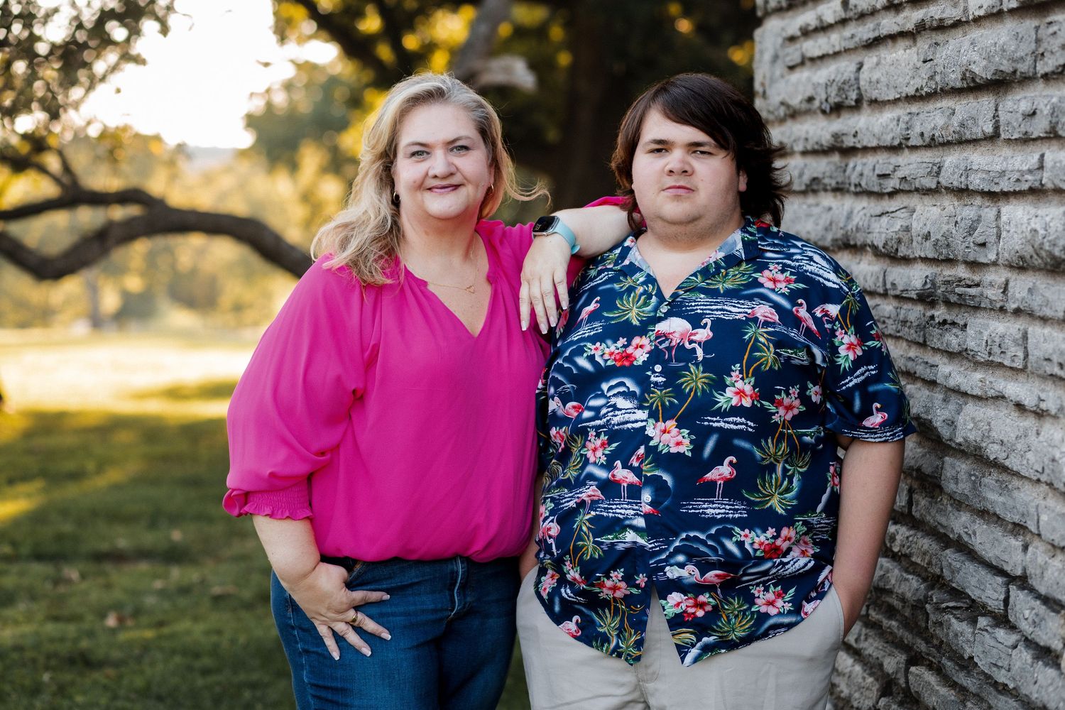 Two people wearing pink and floral print tops pose together against a rustic wooden wall in natural outdoor lighting.