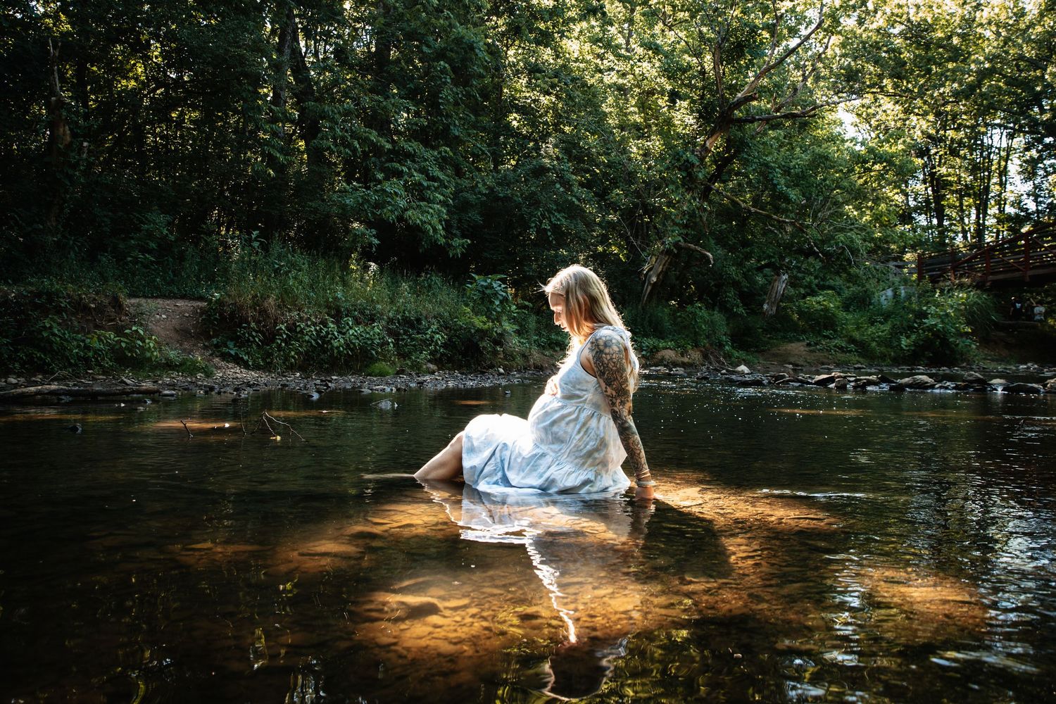 A person in a white dress sits peacefully on a rock in a sunlit forest stream, creating a dreamy atmosphere.