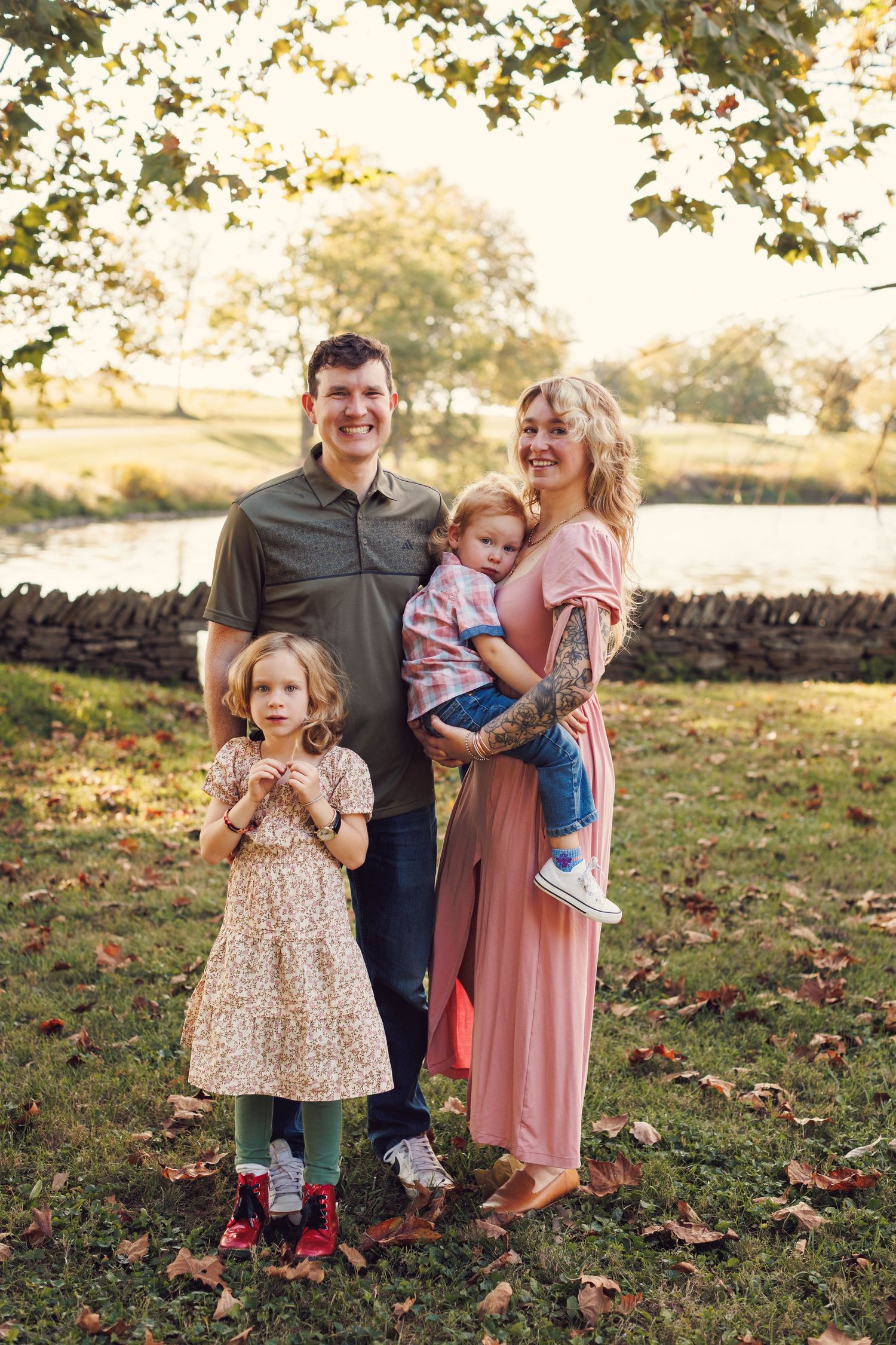 A family stands together on an autumn day with fallen leaves and a stone wall in the background.