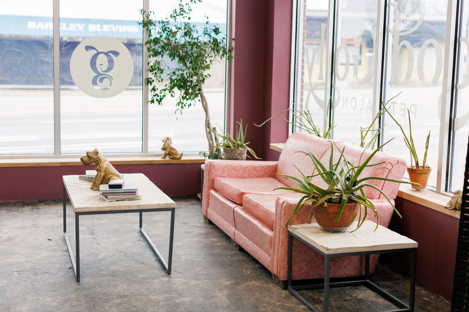 Modern living room with pink velvet sofa, plants and side tables near large windows.