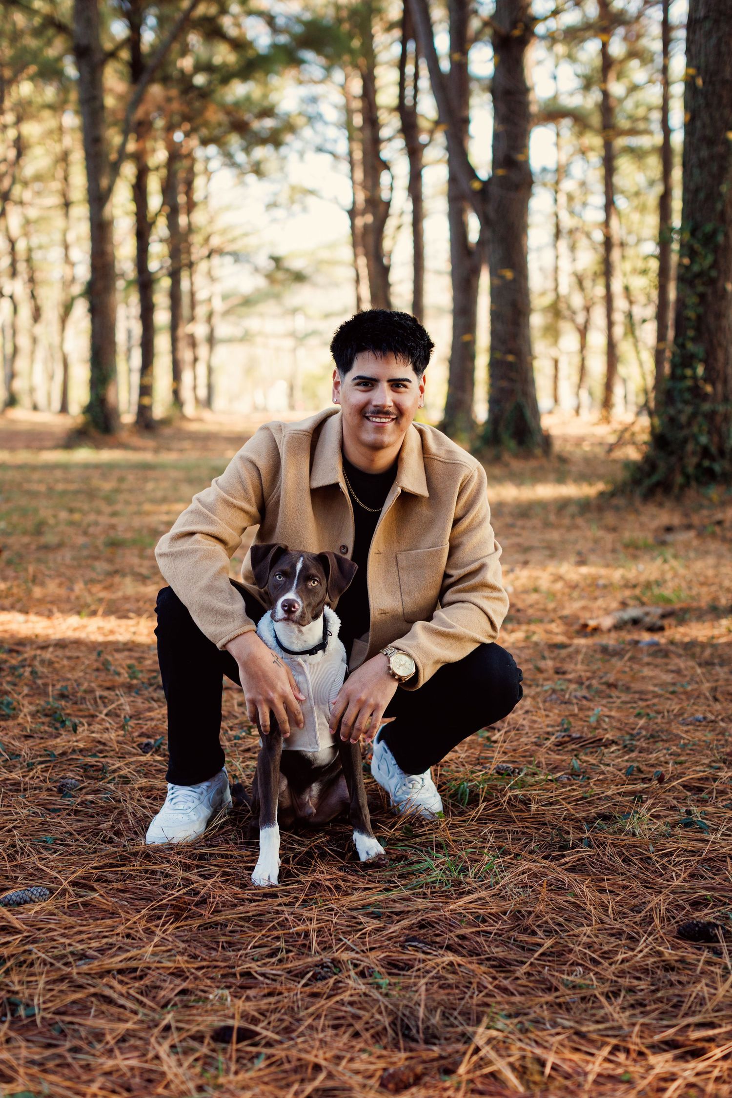 Person in tan jacket crouches with black and white dog on pine needle covered forest floor at sunset.
