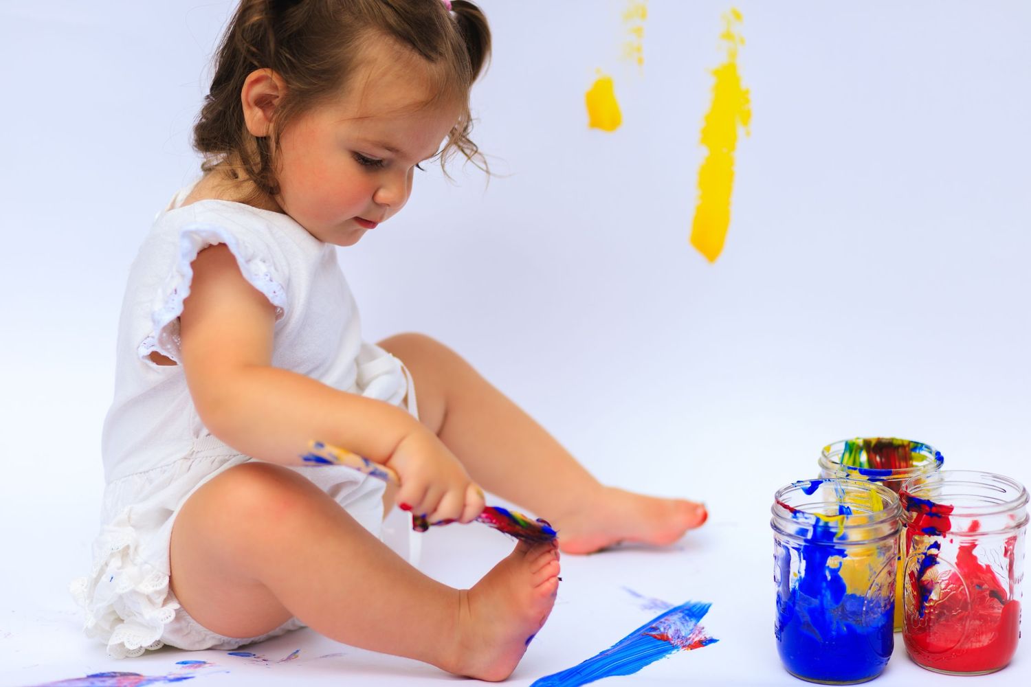 A small child in white clothing creates colorful artwork with paint.