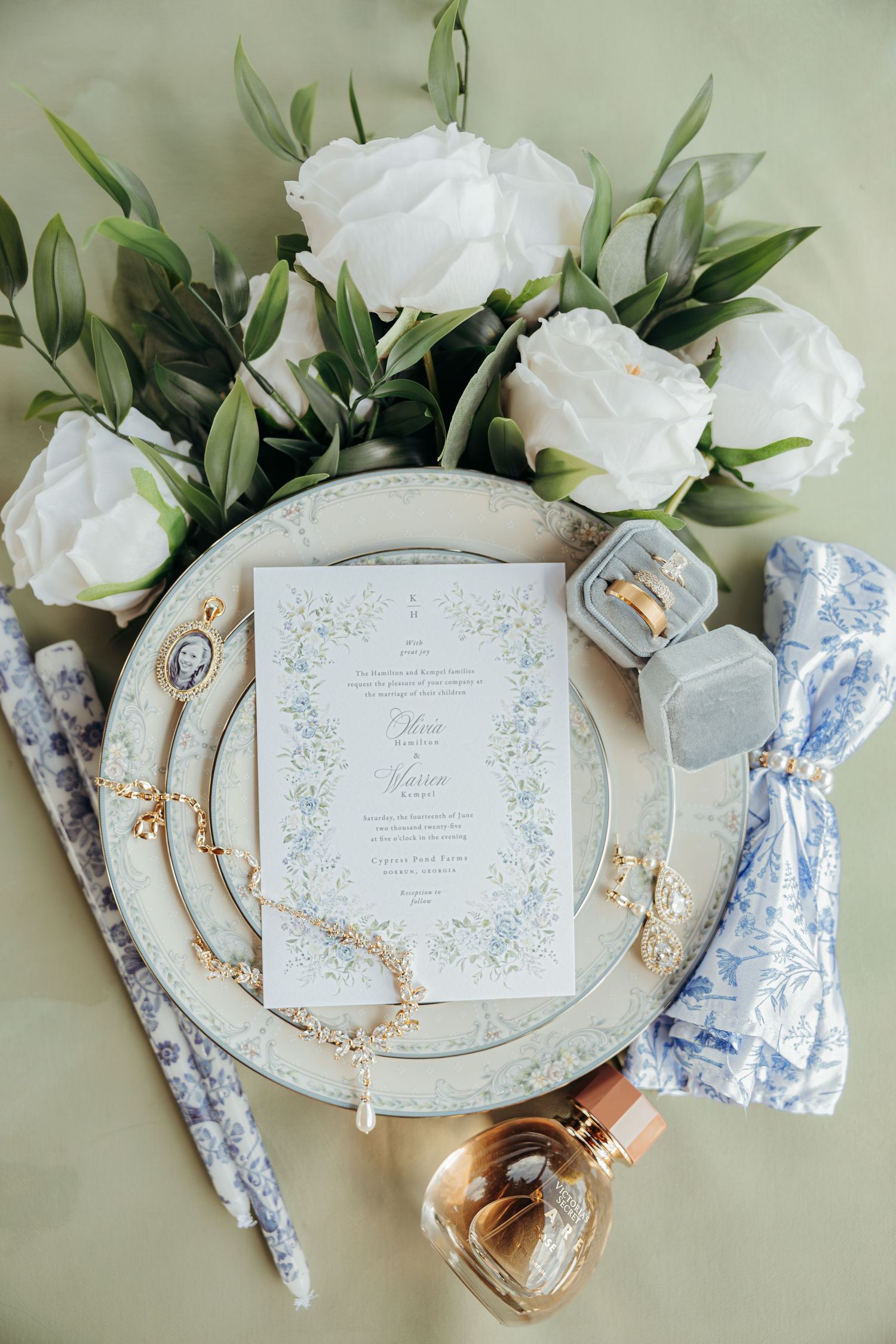 Elegant wedding invitation suite with white roses, blue ribbons, and gold accents arranged on a light beige background.