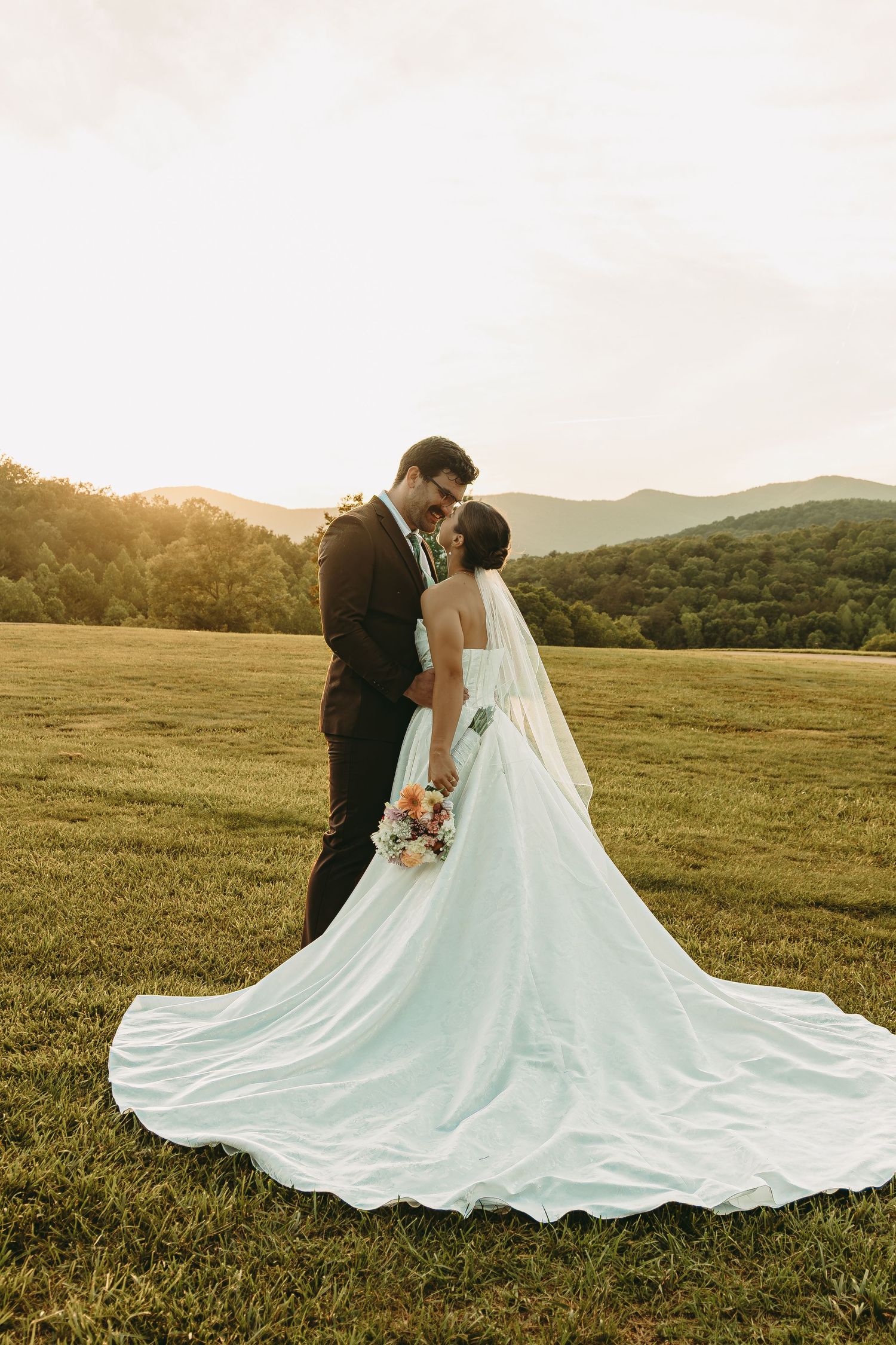 A wedding couple poses in a field at sunset with mountains in the background and flowing white dress.