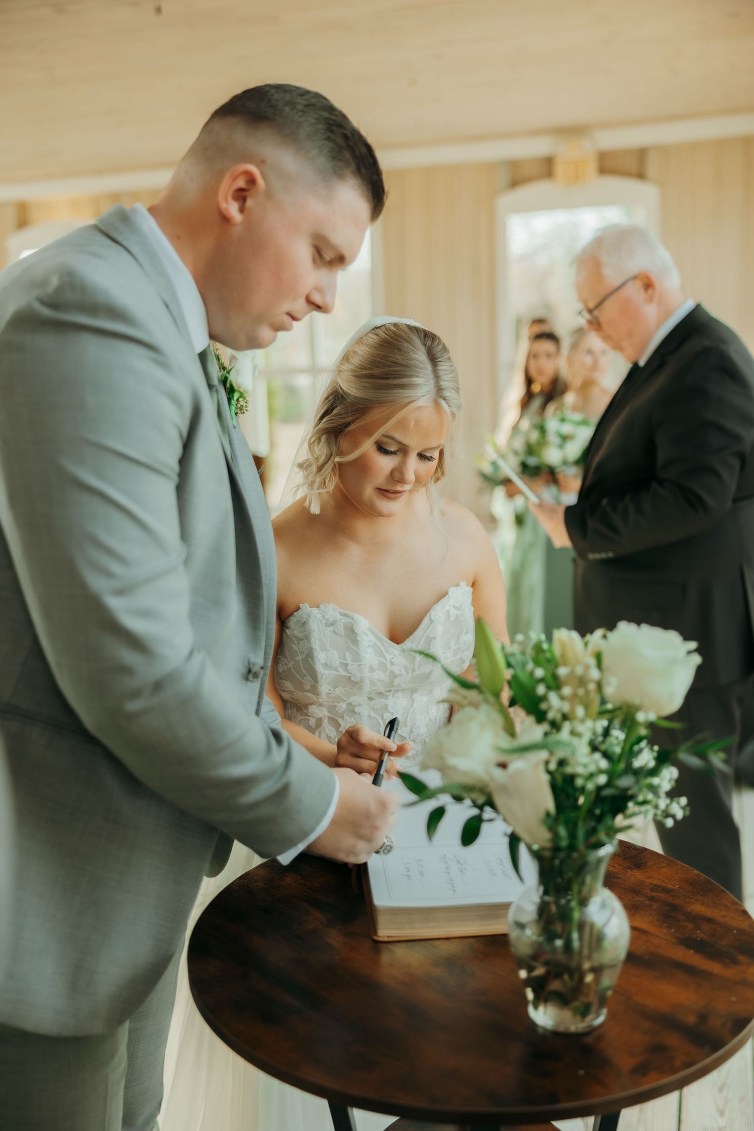 A couple in formal wedding attire signs documents at a wooden table with floral decor during their ceremony.