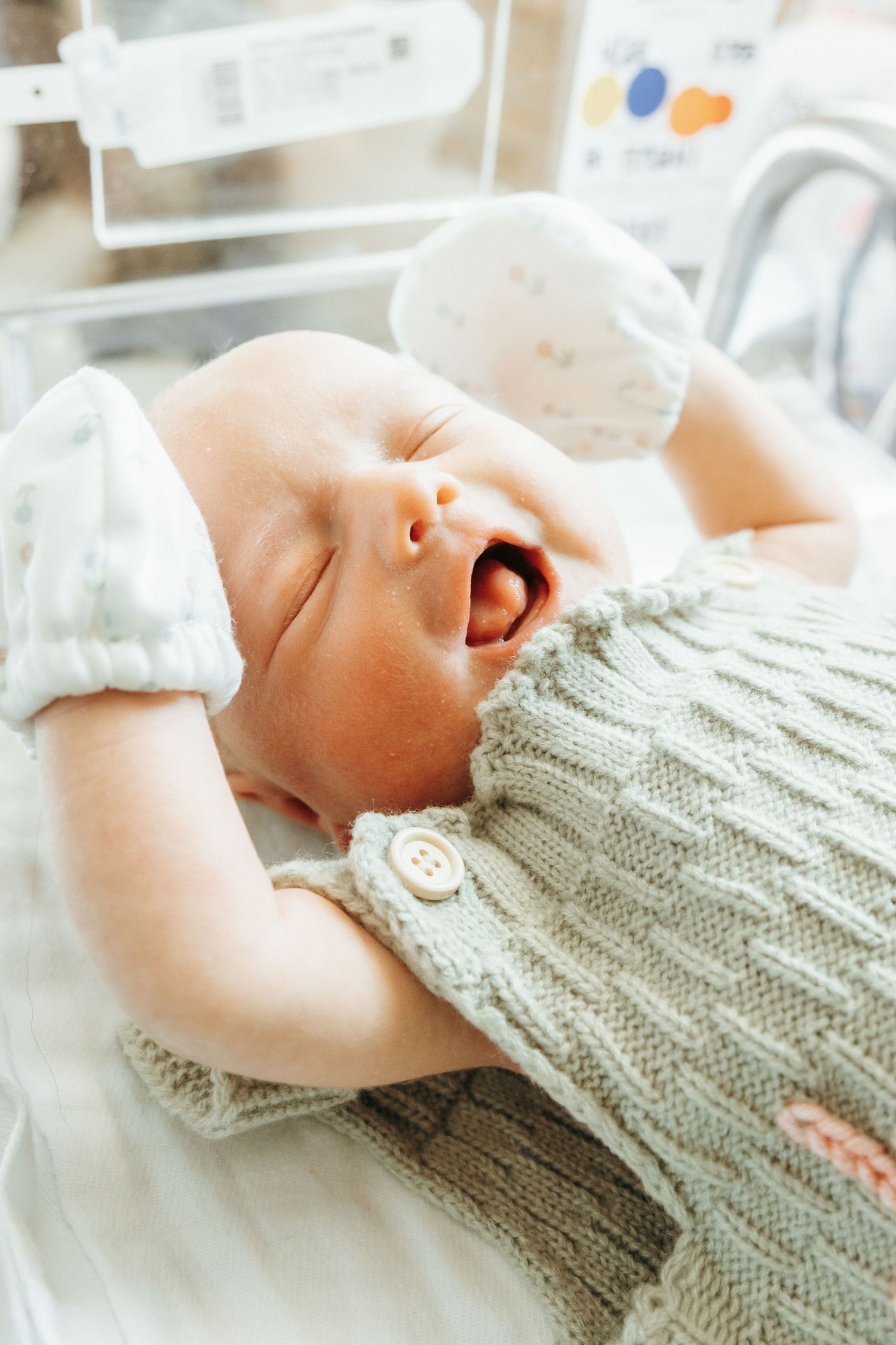 Close up view of an infant wearing a cozy gray knit sweater while lying on soft white bedding.