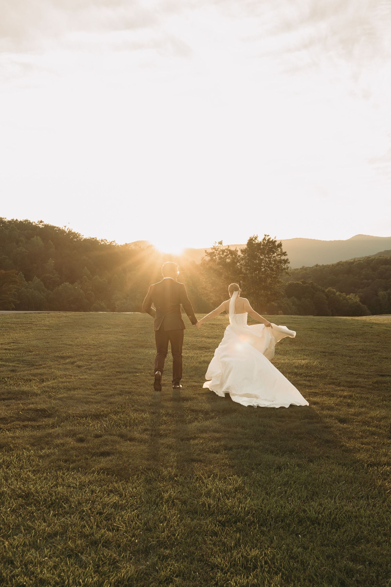 Bride and groom share a playful moment while walking through a sunlit field at dusk.