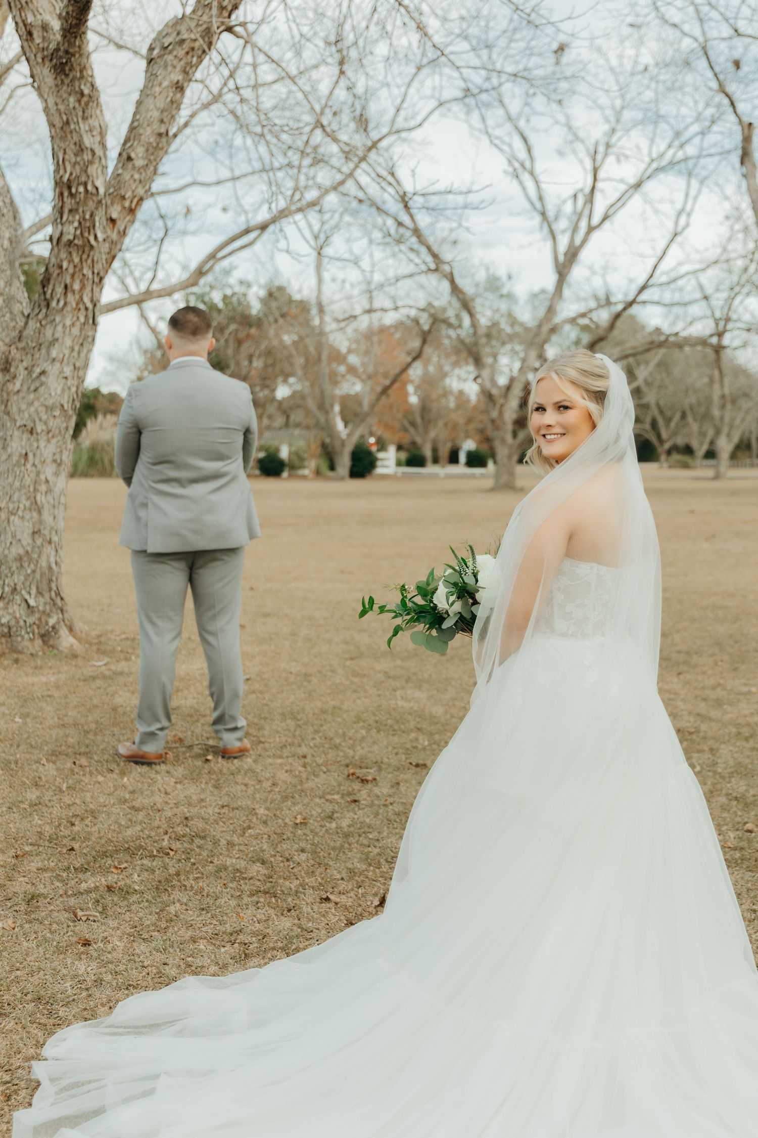 A bride in a flowing white gown stands behind a groom in a grey suit in an outdoor field with bare trees.