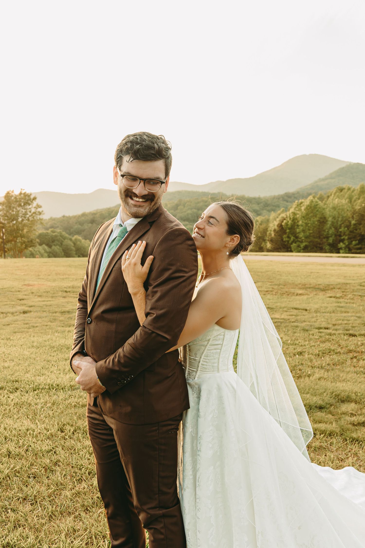 A couple shares a joyful moment in their wedding attire against a backdrop of rolling hills during golden hour.