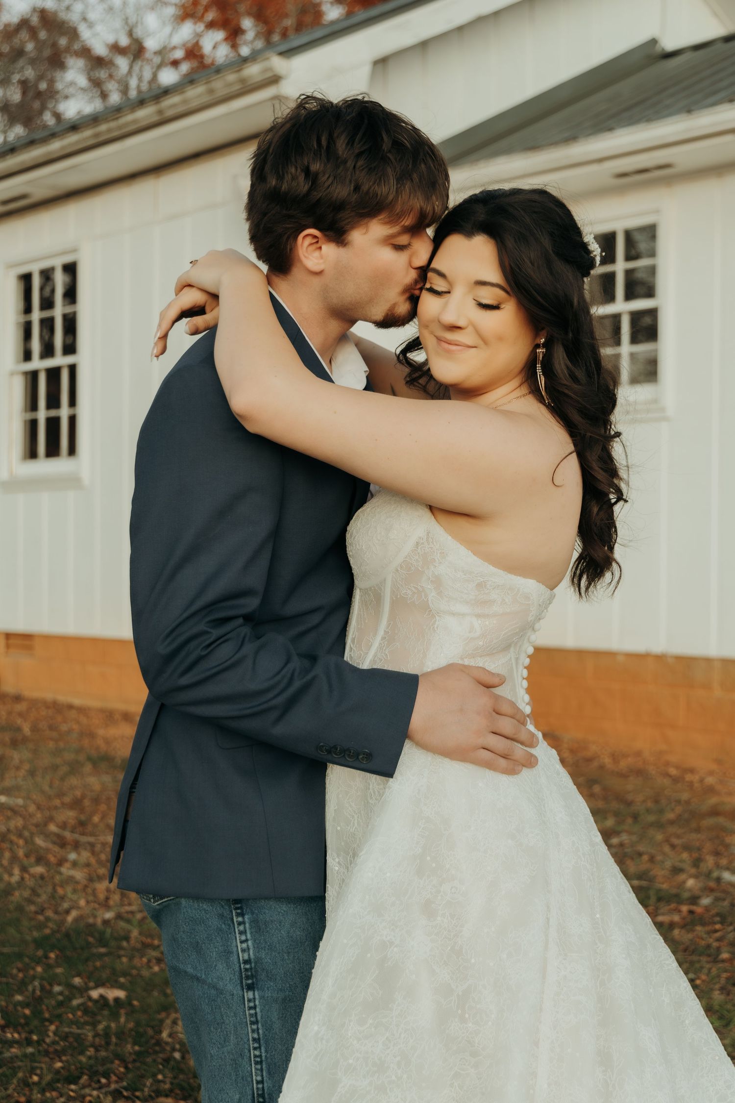 A romantic wedding couple embraces outdoors in autumn with a white building in the background.