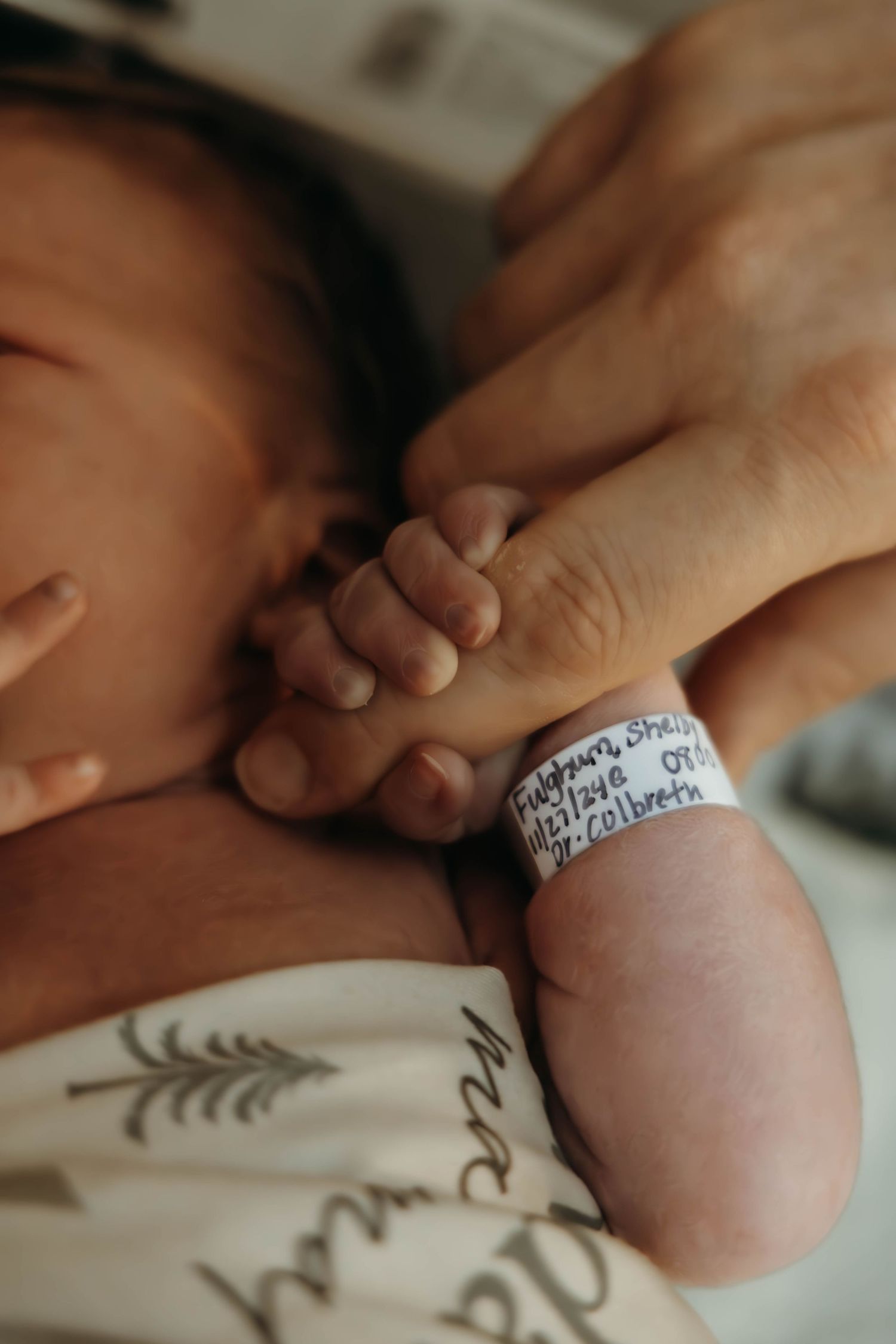 Close up of newborn baby wearing hospital ID bracelet holding an adult finger with white sheet in background.