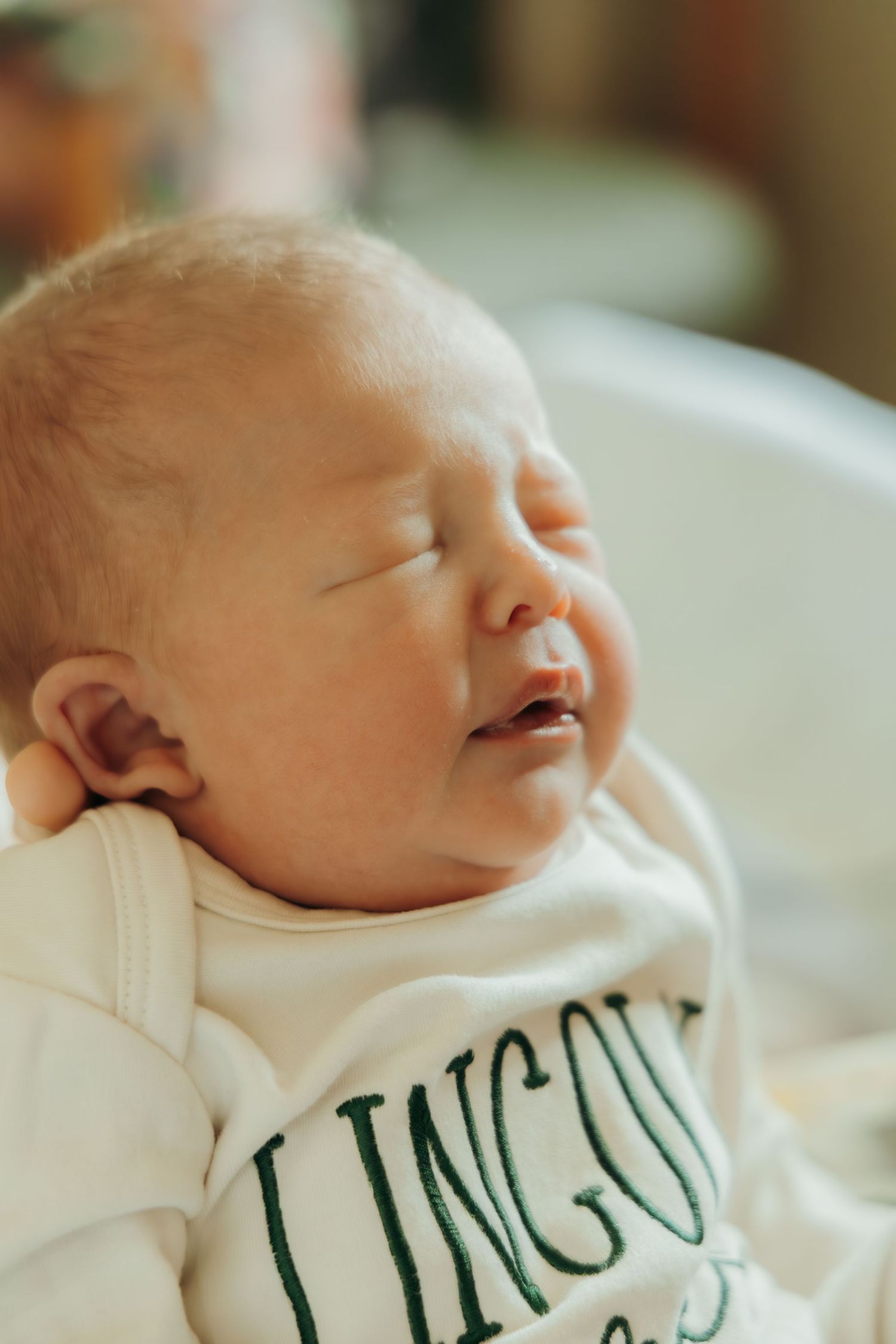 A newborn baby sleeps peacefully while wearing a white onesie with green text.