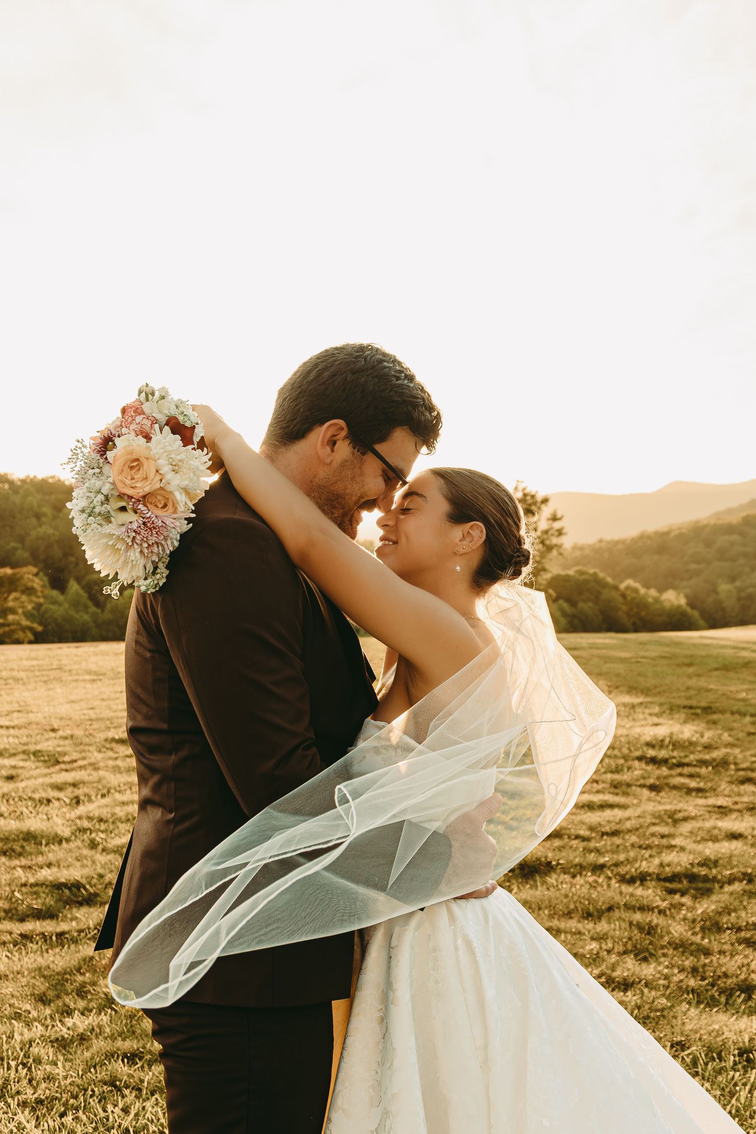 Newlywed couple embracing in a grassy field during golden hour with flowing white wedding veil.