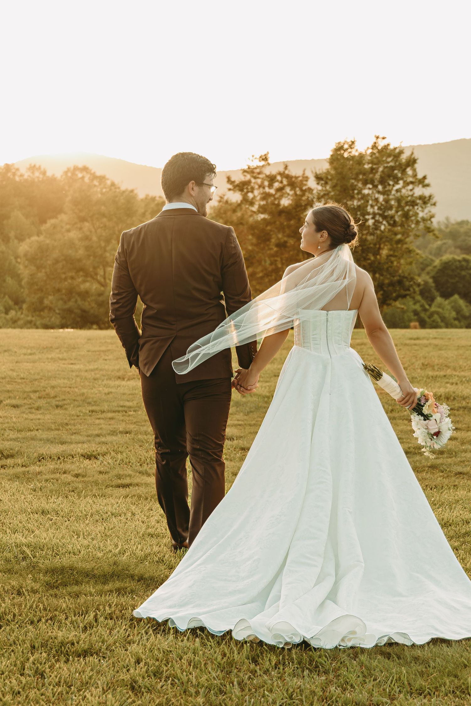 A couple in wedding attire walks through a sunset-lit field with mountains in the distance.