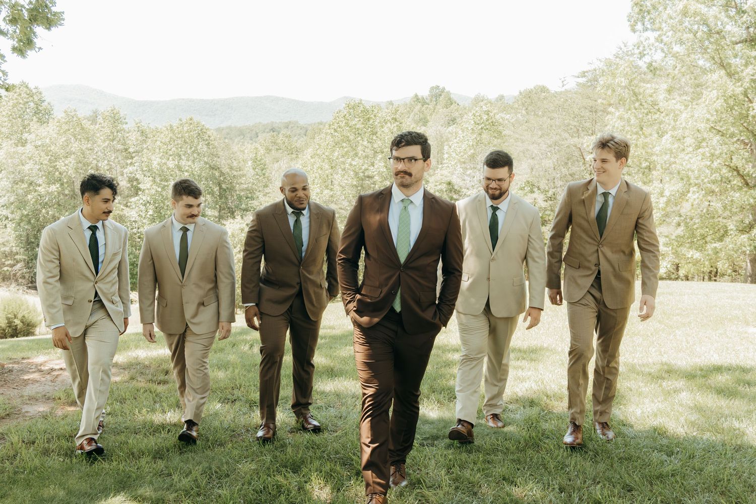 Groomsmen in coordinated suits walking together across sunny hillside.