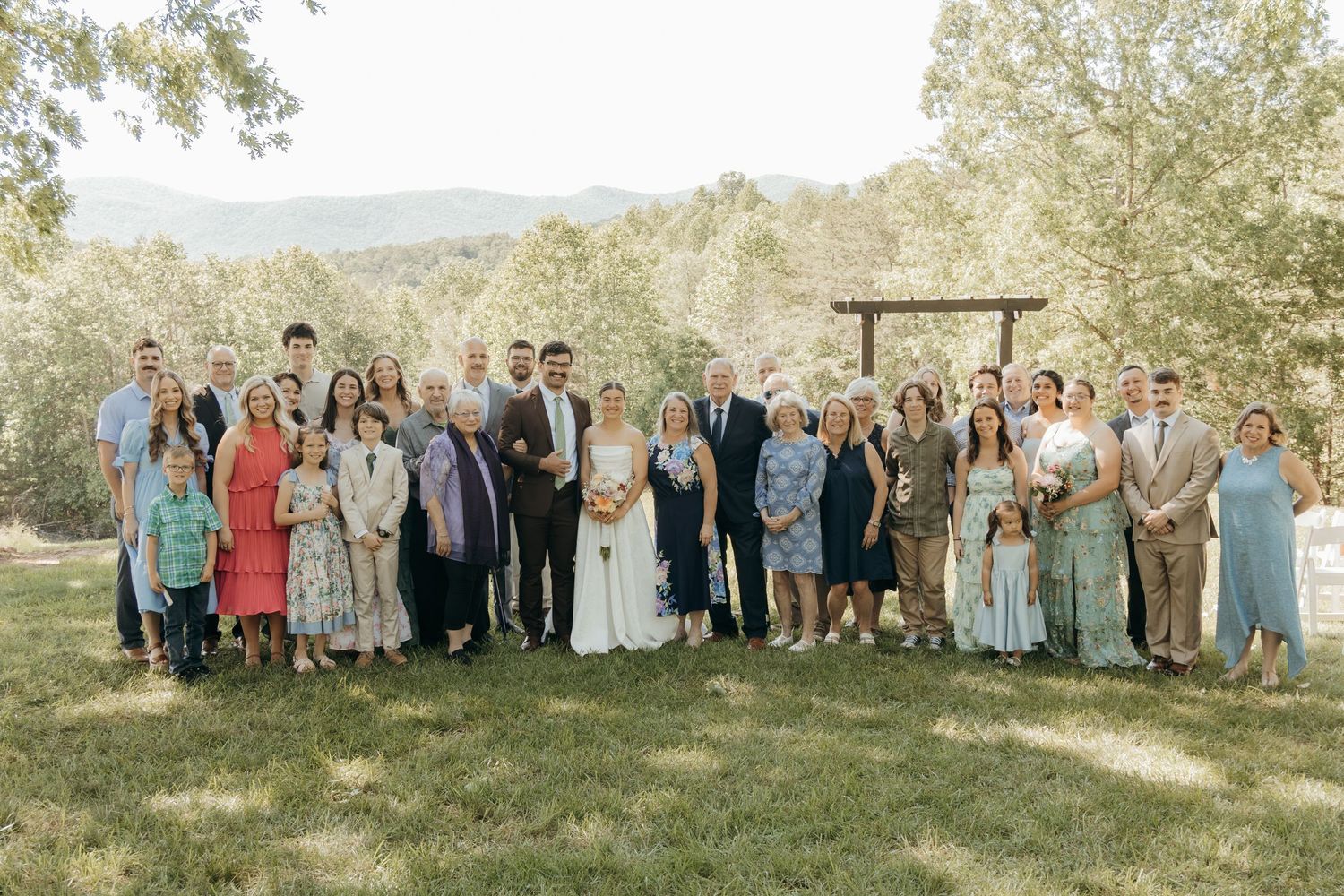 Wedding party and guests gather for a group photo on a grassy hillside with mountain views.