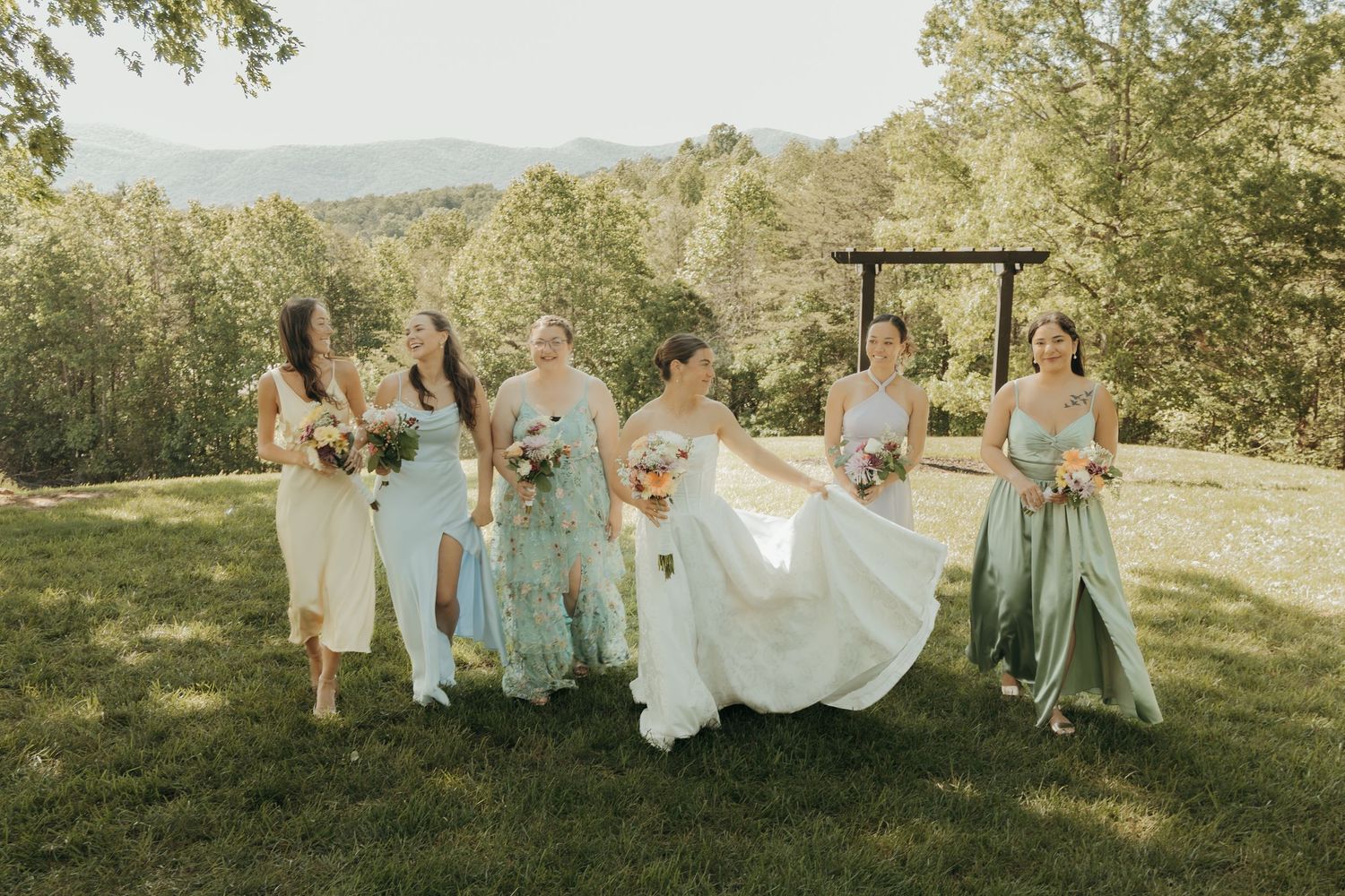 Bridesmaids in mint green dresses laugh while walking with bride on grassy hillside.