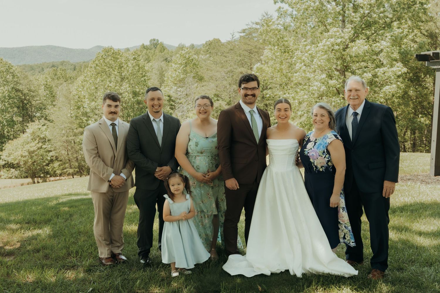Family members gather for a formal portrait at an outdoor wedding with rolling hills behind them.