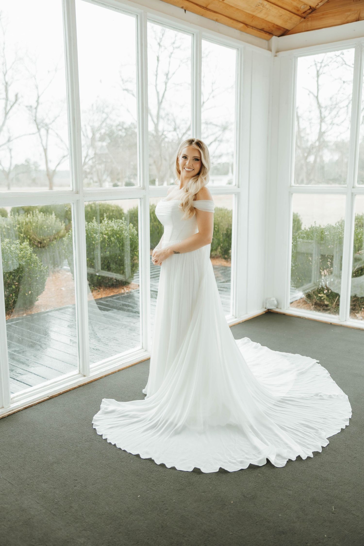 A flowing white wedding dress with a long train twirls elegantly in a bright sunlit room with wood ceiling and windows.