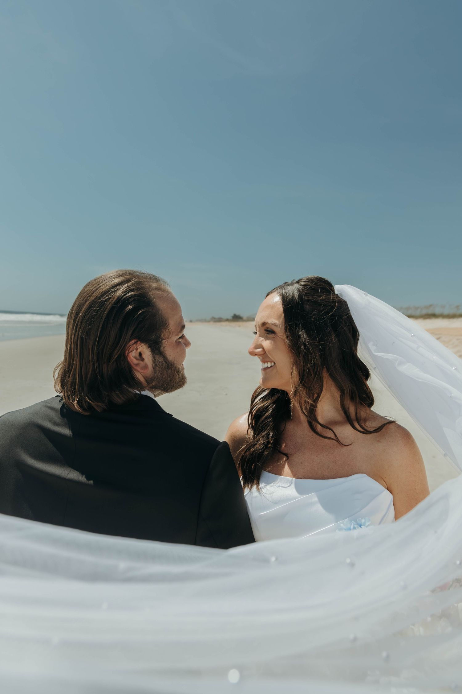 A romantic wedding moment captured on a beach as a flowing white veil wraps around the newlyweds.