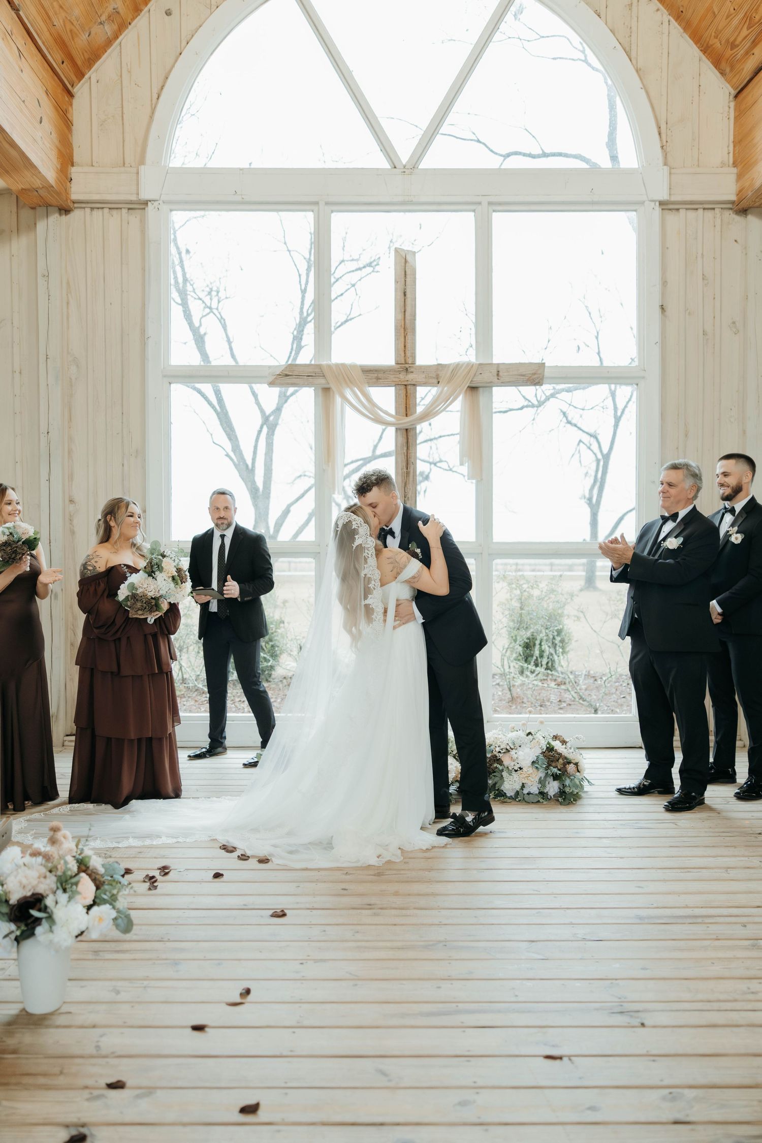 A wedding ceremony takes place in a rustic chapel with wooden floors and large windows, decorated with floral arrangements.