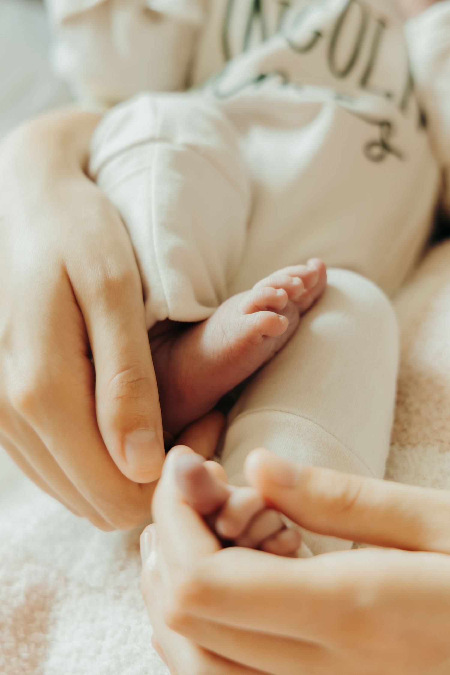 A gentle hand tenderly holds a tiny baby foot against a soft white background in warm natural lighting.