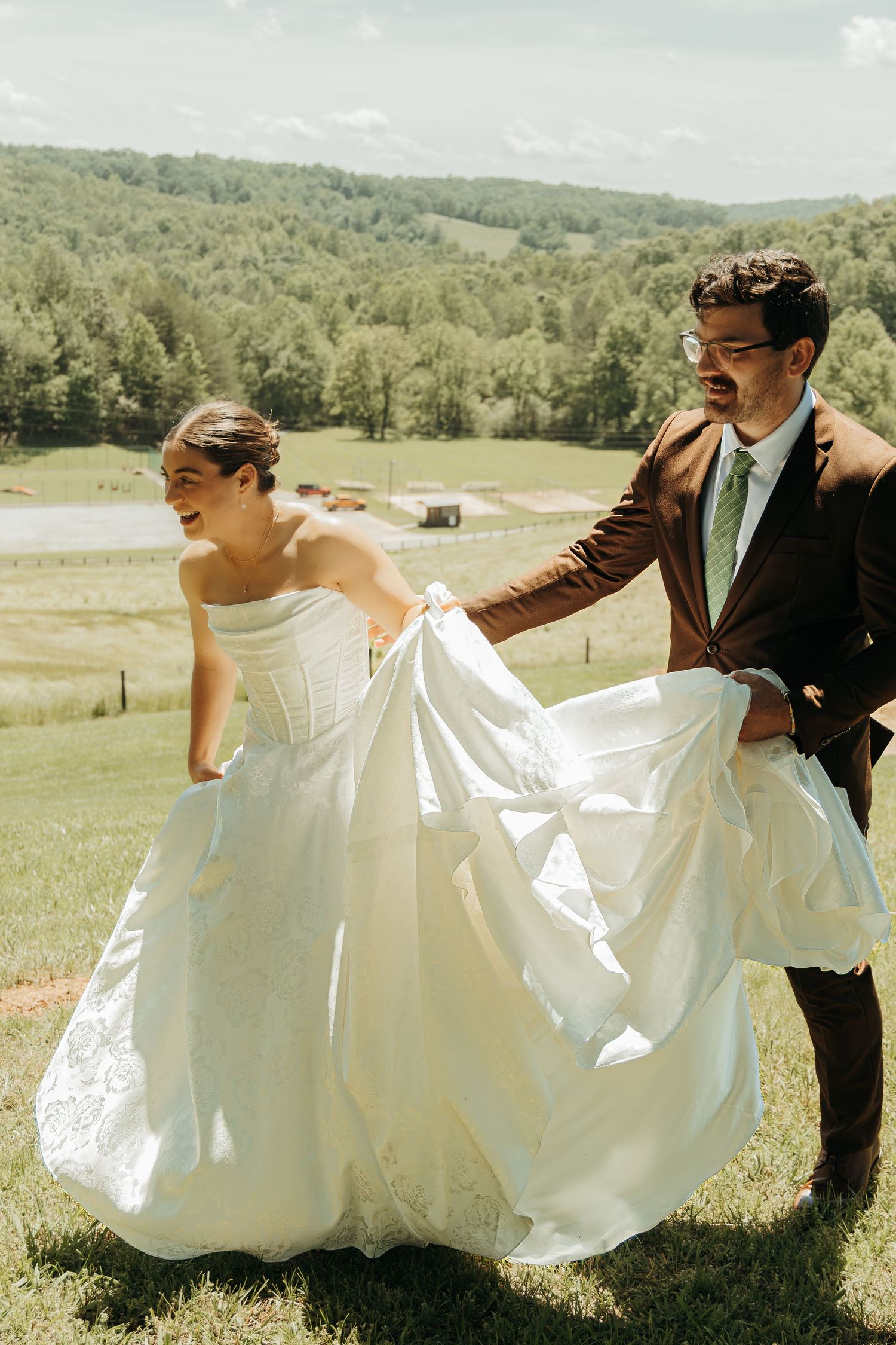Wedding couple in formal attire walking through scenic countryside with flowing dress.