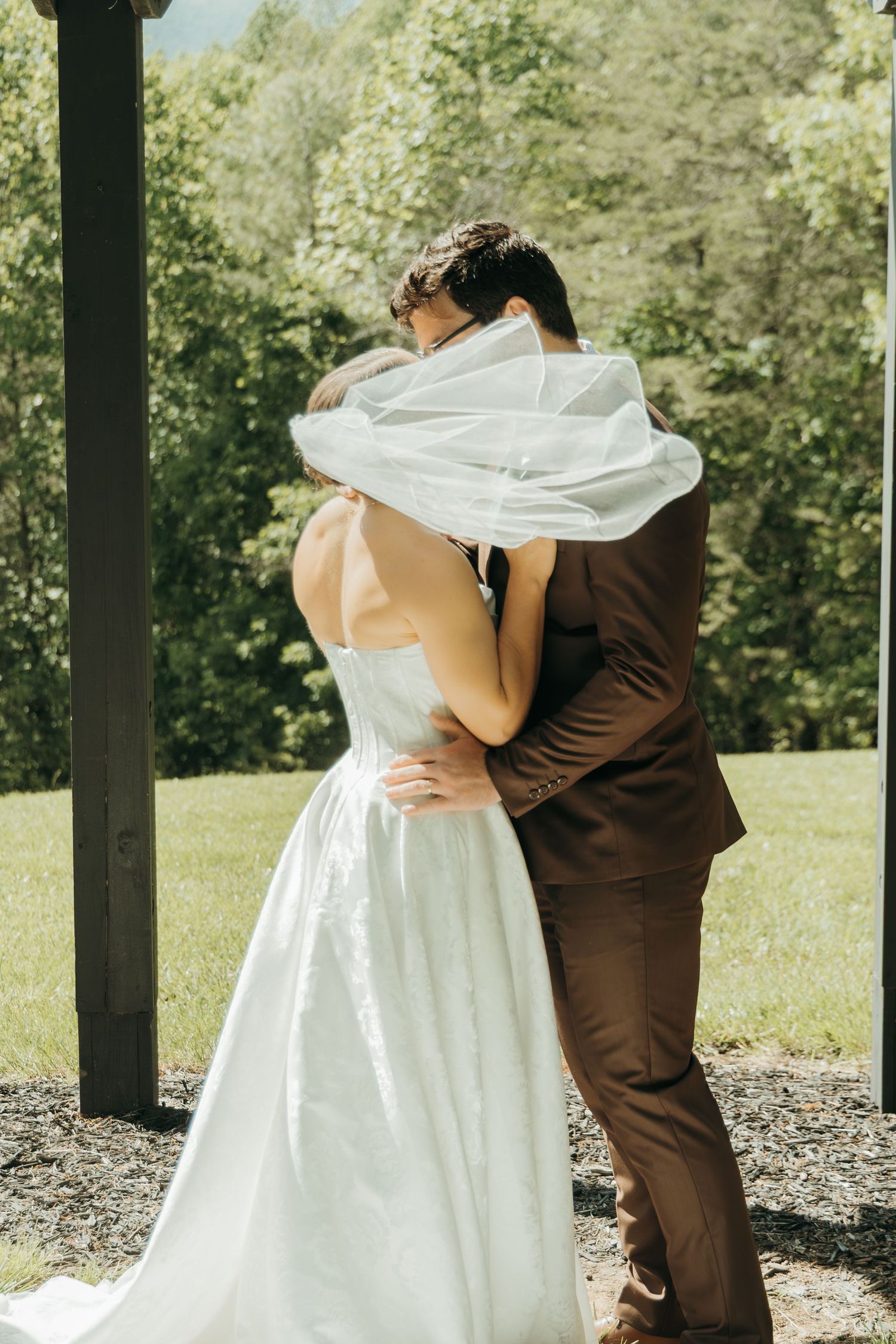 Romantic first kiss moment under wooden pergola with bride's veil flowing in the wind.
