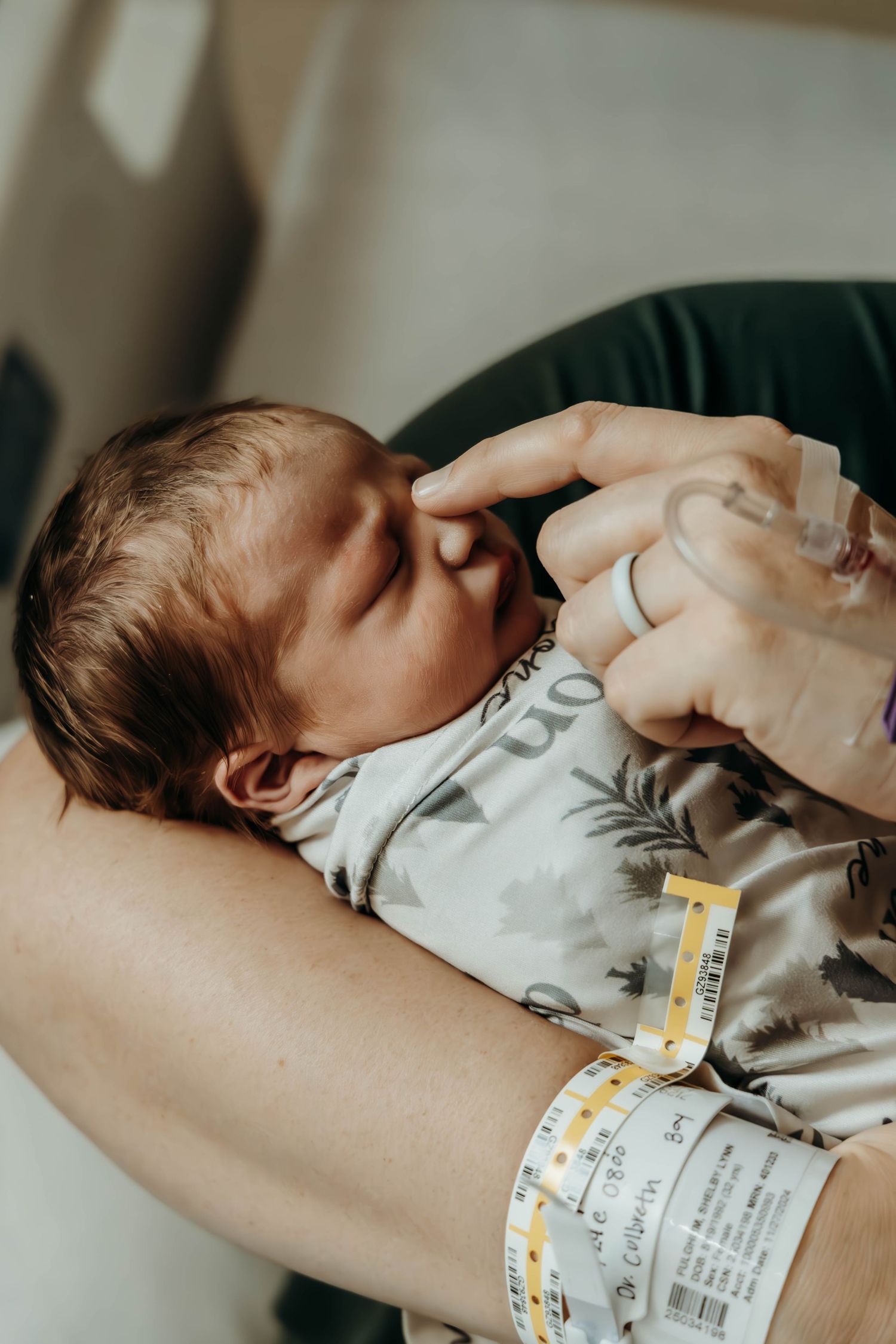A newborn baby wearing a white onesie lies in someone's arms with a hospital ID bracelet visible.