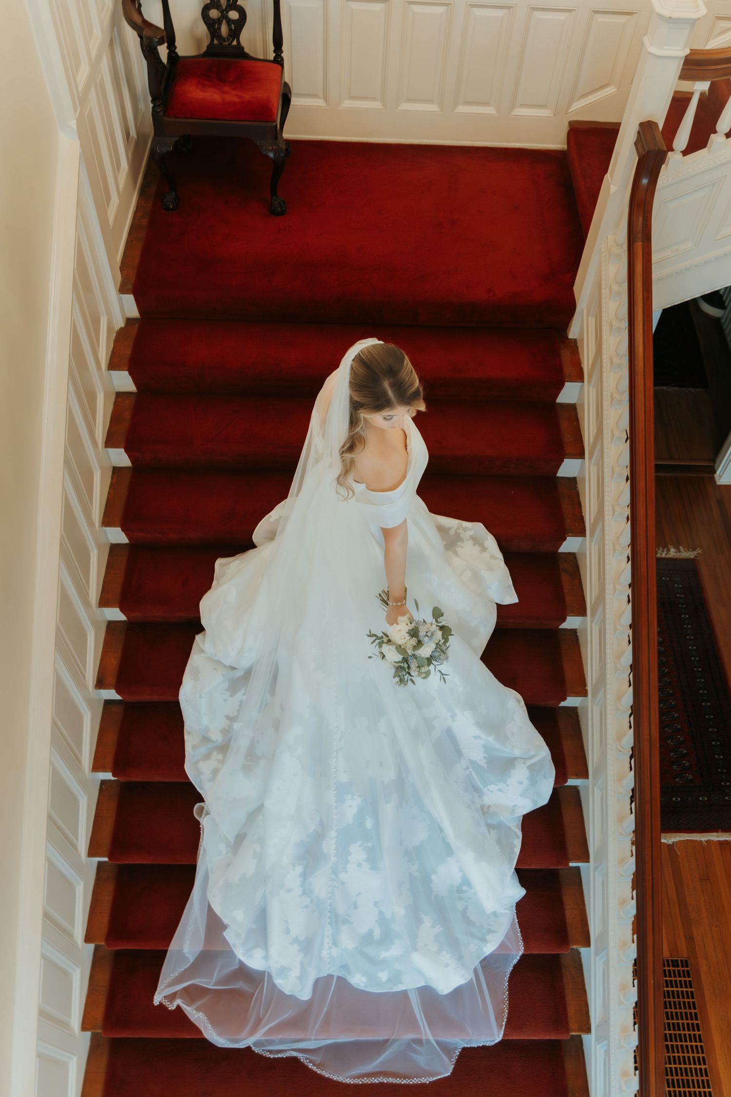 A bride in a flowing white wedding gown stands on red carpeted stairs, photographed from above.