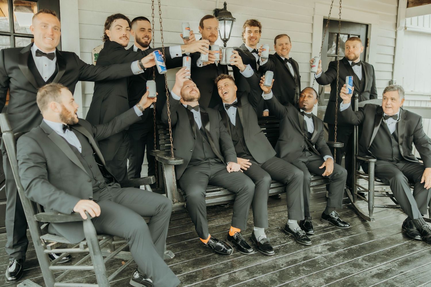 Wedding party in black suits sit casually on outdoor porch while laughing and enjoying candid moment together.