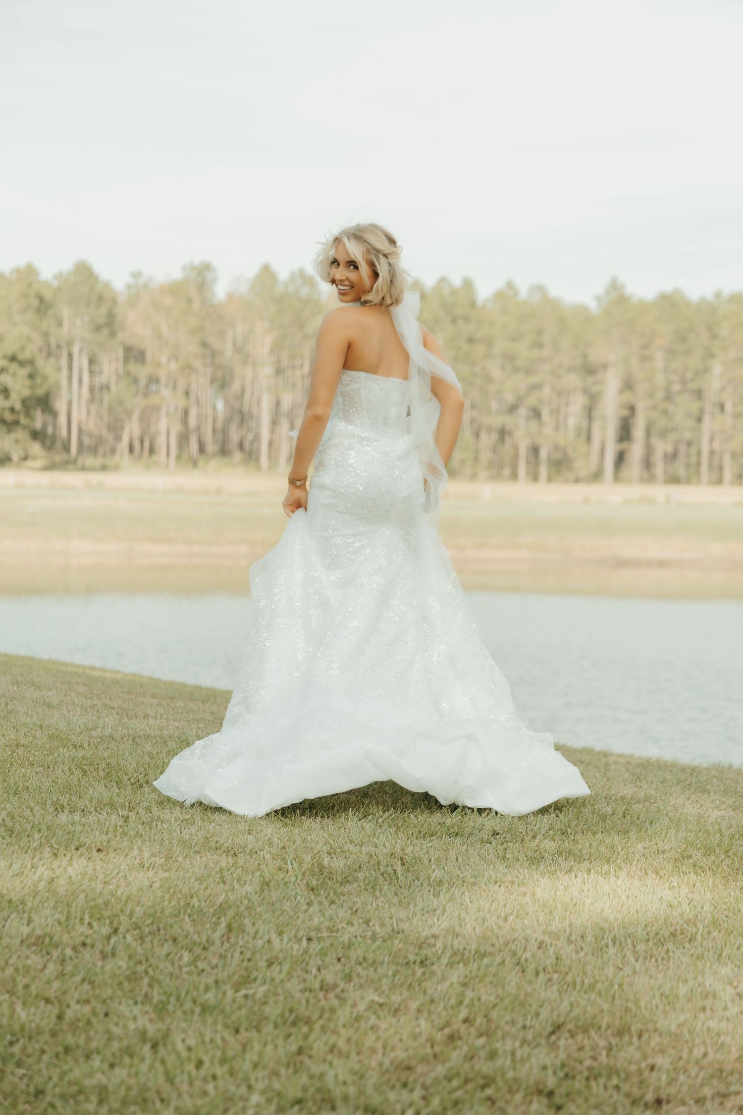 A flowing white wedding dress swirls gracefully across green grass near a serene lake with trees in the background.