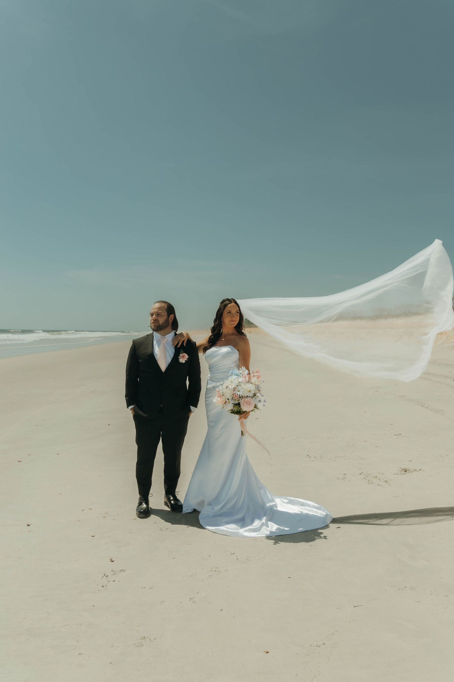 A couple in wedding attire poses on white sand dunes with a flowing veil caught in the wind against a blue sky.