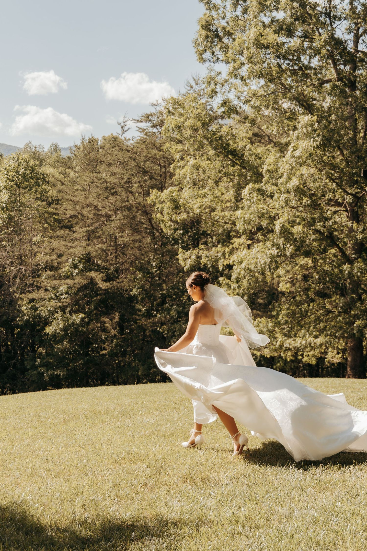 Bride in flowing white dress twirls on grassy hillside with mountains in background.