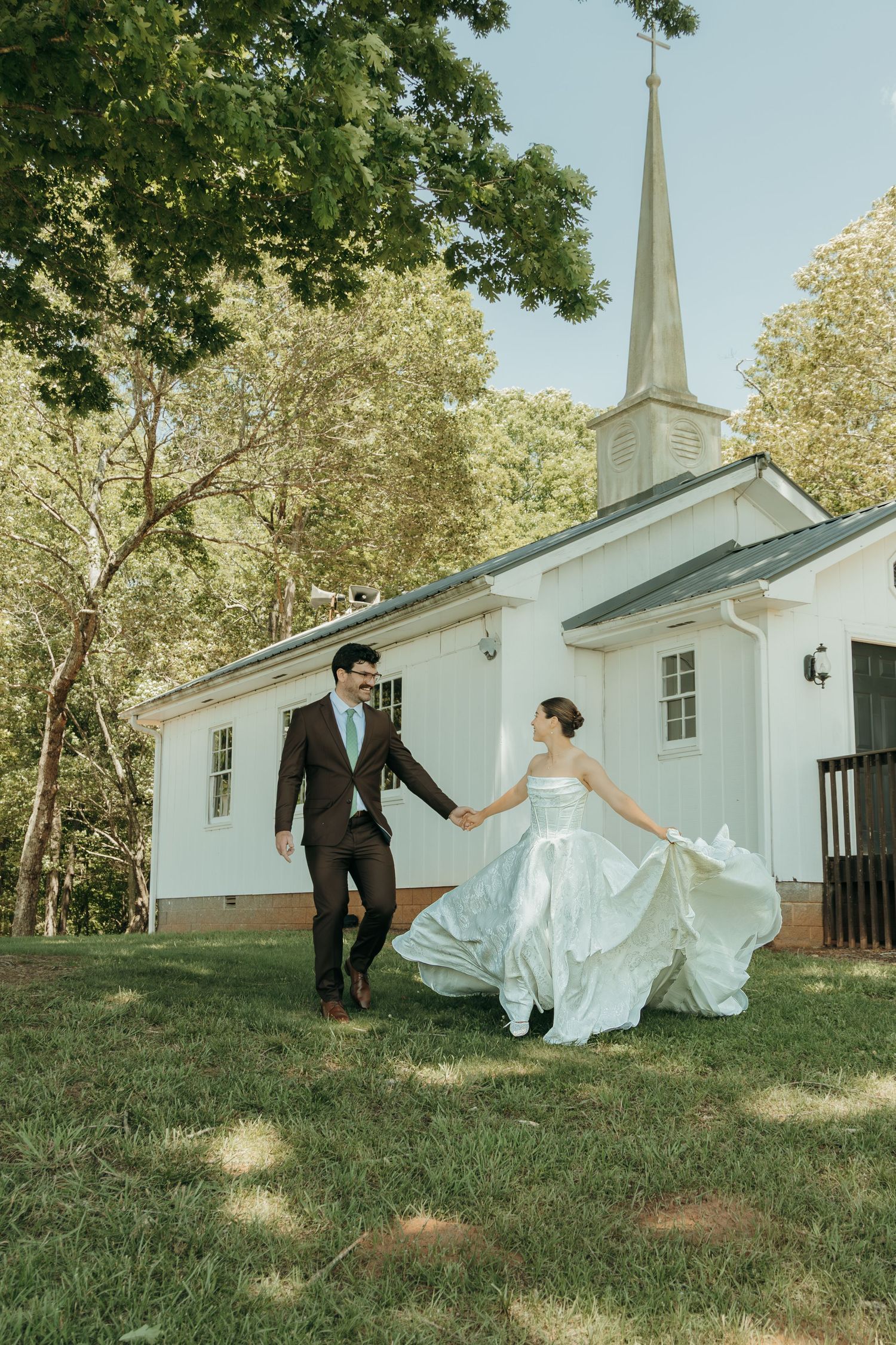 A couple runs playfully in front of a white chapel with a tall steeple on their wedding day.