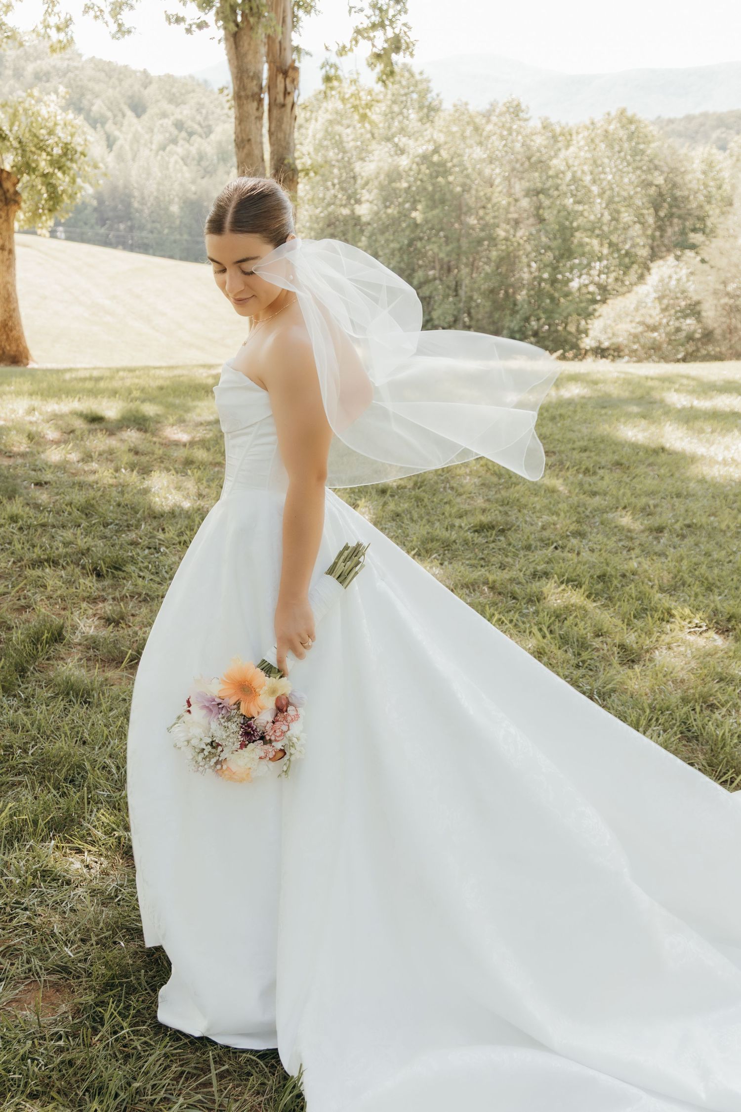 Elegant bride in flowing white gown poses with bouquet on sunny hillside.